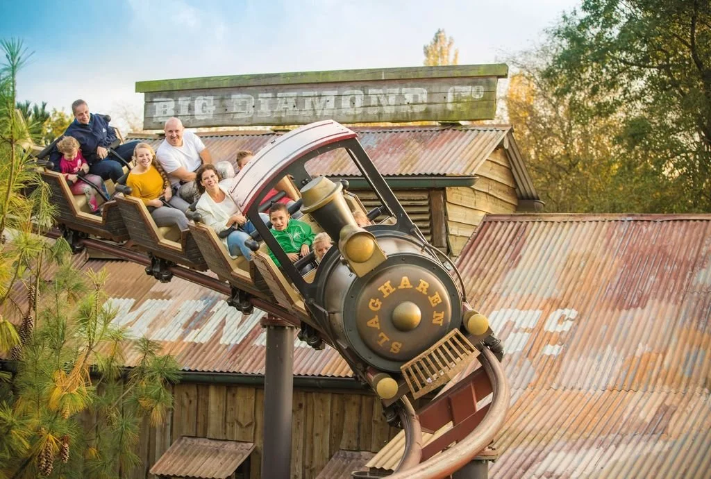 A family riding a vintage-style roller coaster at an amusement park with autumn trees in the background.