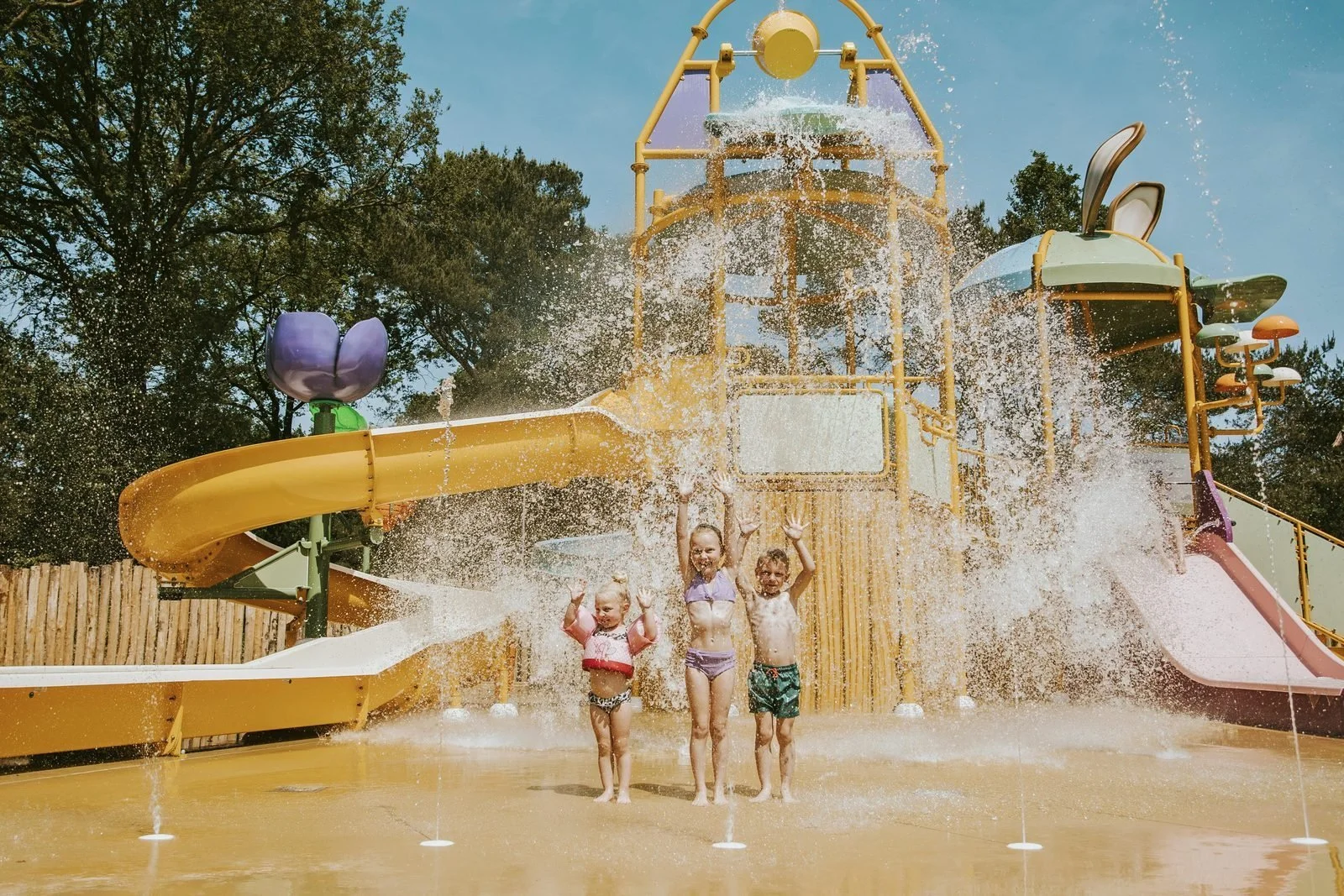 Three children playing on a water playground with slides and water spray under clear blue sky.
