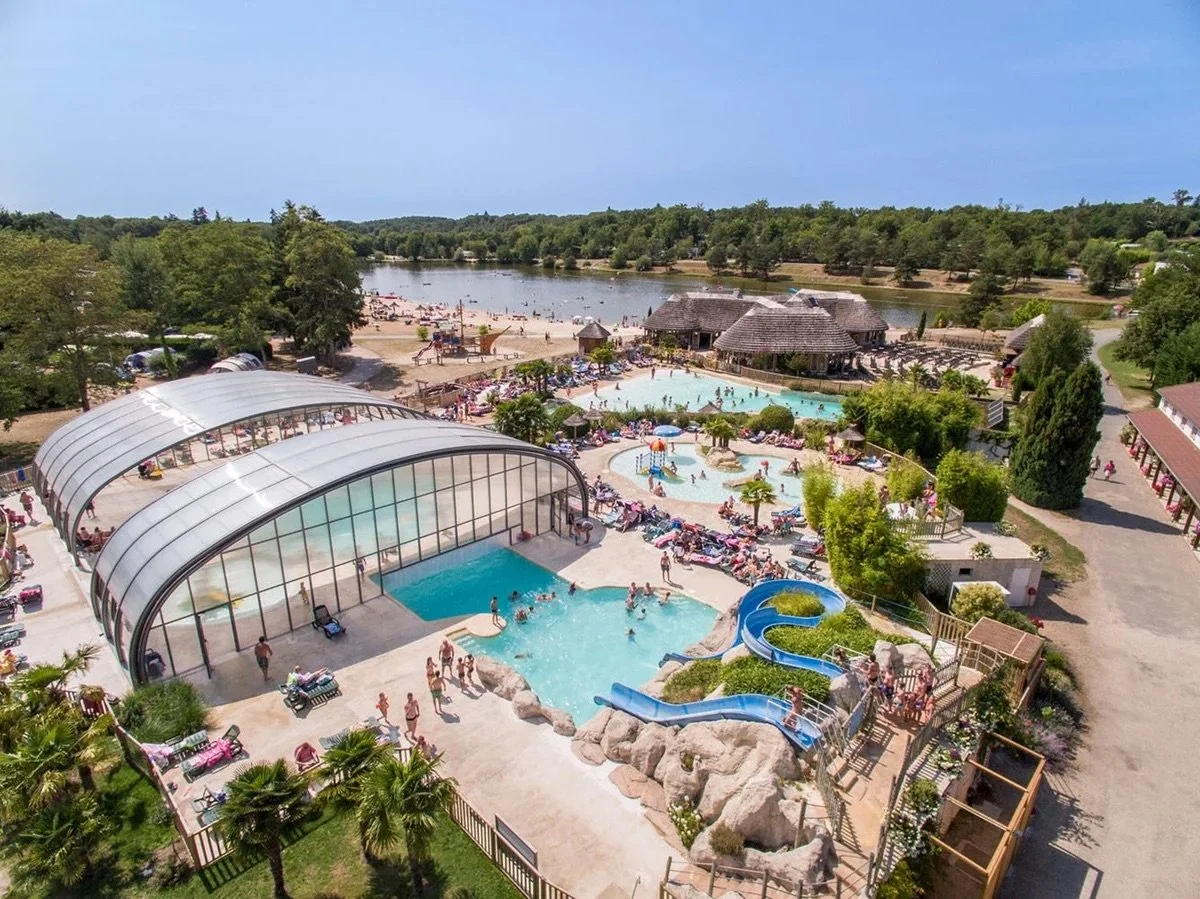 Aerial view of a large outdoor water park with multiple pools, waterslides, and a lazy river, surrounded by trees and a beach area near a lake.