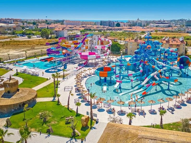 Aerial view of a large water park with multiple colorful waterslides, a lazy river, and lounging areas, with buildings and the ocean in the background.