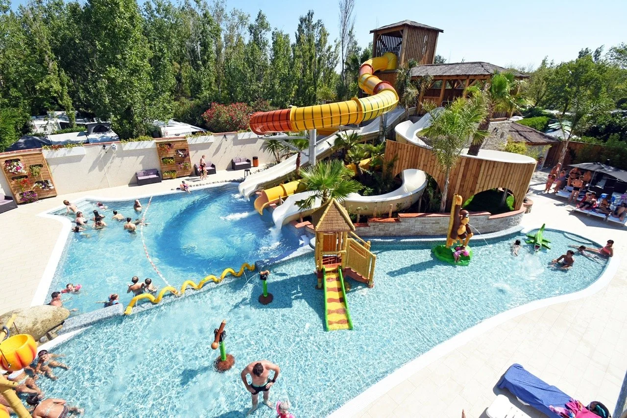 People enjoying a water park pool with water slides, tropical plants, and lounge chairs.