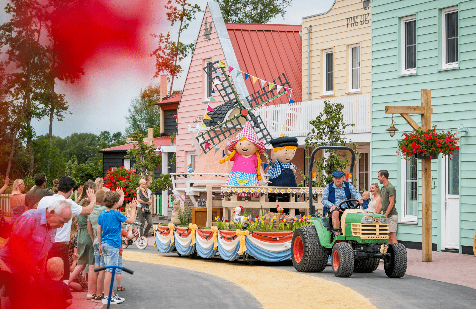 Kleurrijke praalwagen met vrolijke figuren en een molen die door de straat rijdt tijdens een feestelijke optocht, omringd door toeschouwers.
