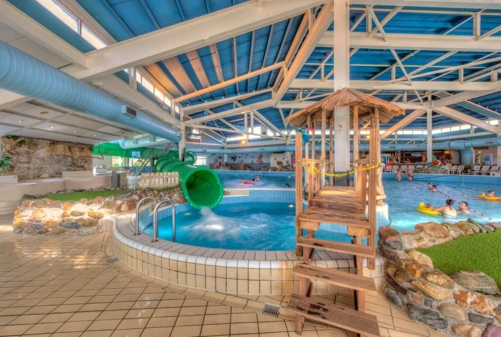 Indoor water park with a large swimming pool, a green water slide, a thatched-roof platform, and people swimming and playing in the water.