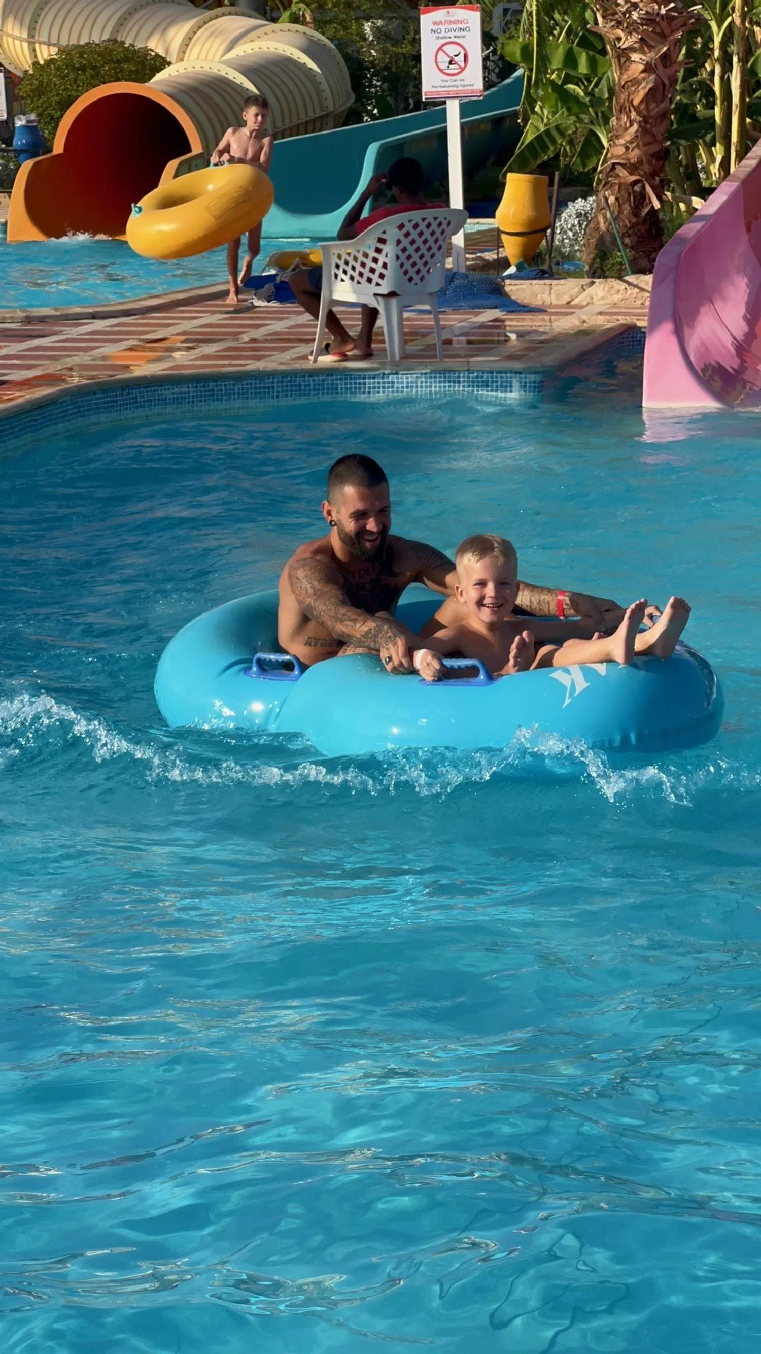 Een man en een jonge jongen zitten samen op een blauwe opblaasband in het zwembad. Ze lachen en genieten van hun tijd samen op een zonnige dag.