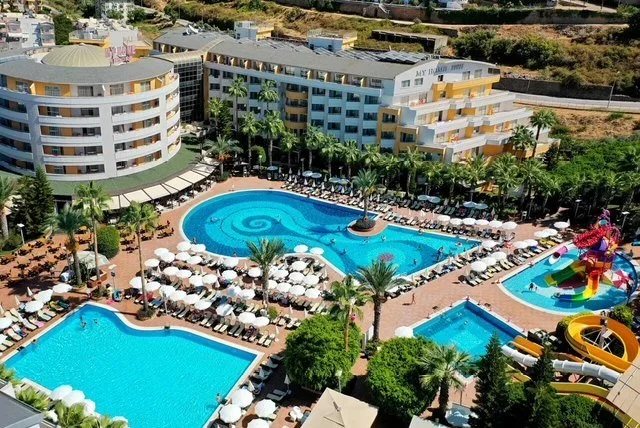 Aerial view of a hotel pool area with multiple pools, lounge chairs, umbrellas, water slides, and surrounding multi-story hotel buildings with palm trees.