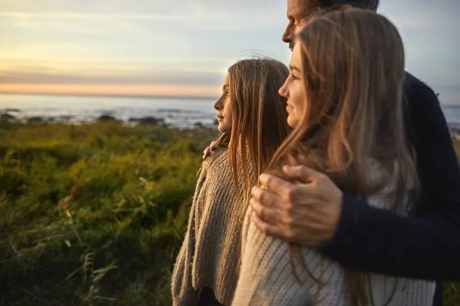 A family of three standing outdoors during sunset, with the father hugging the mother from behind, and the daughter standing between them, all smiling softly.