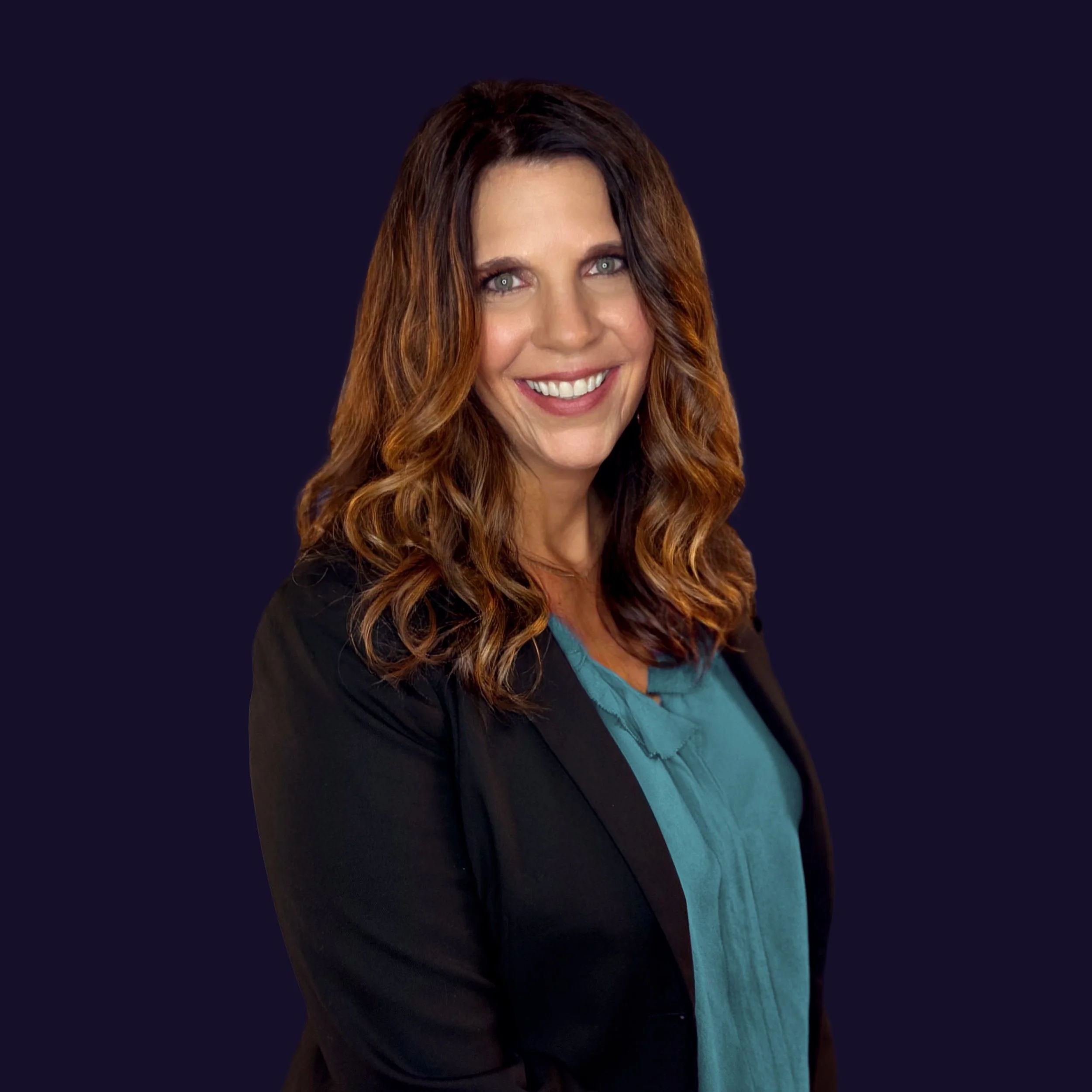 A woman with long, wavy brown hair and blue eyes, wearing a dark blazer over a teal blouse, smiling at the camera against a dark background.