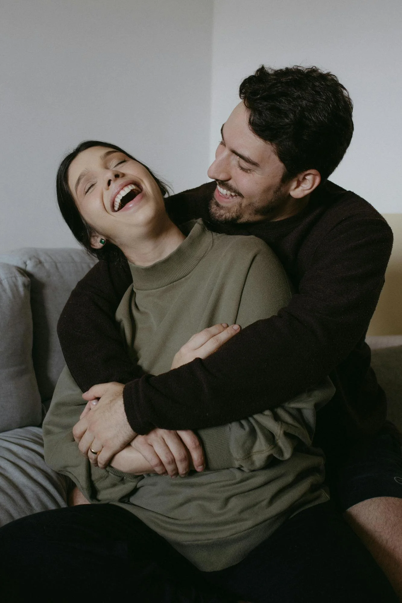 couple embracing and laughing - woman with black hair un bun and olive sweater, man wrapping his arms around her in black sweater and black hair