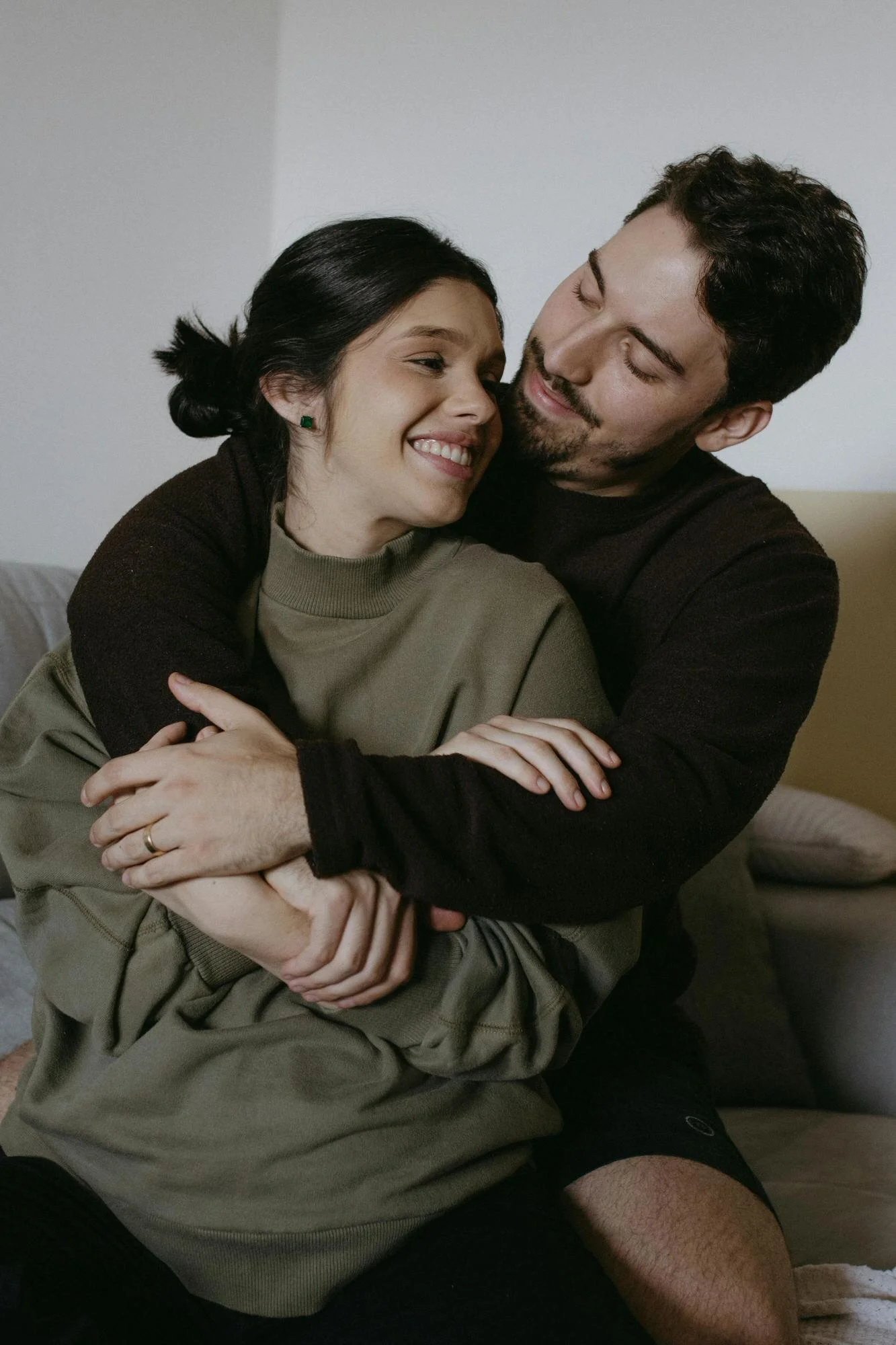 couple embracing and smiling - woman with black hair un bun and olive sweater, man wrapping his arms around her in black sweater and black hair