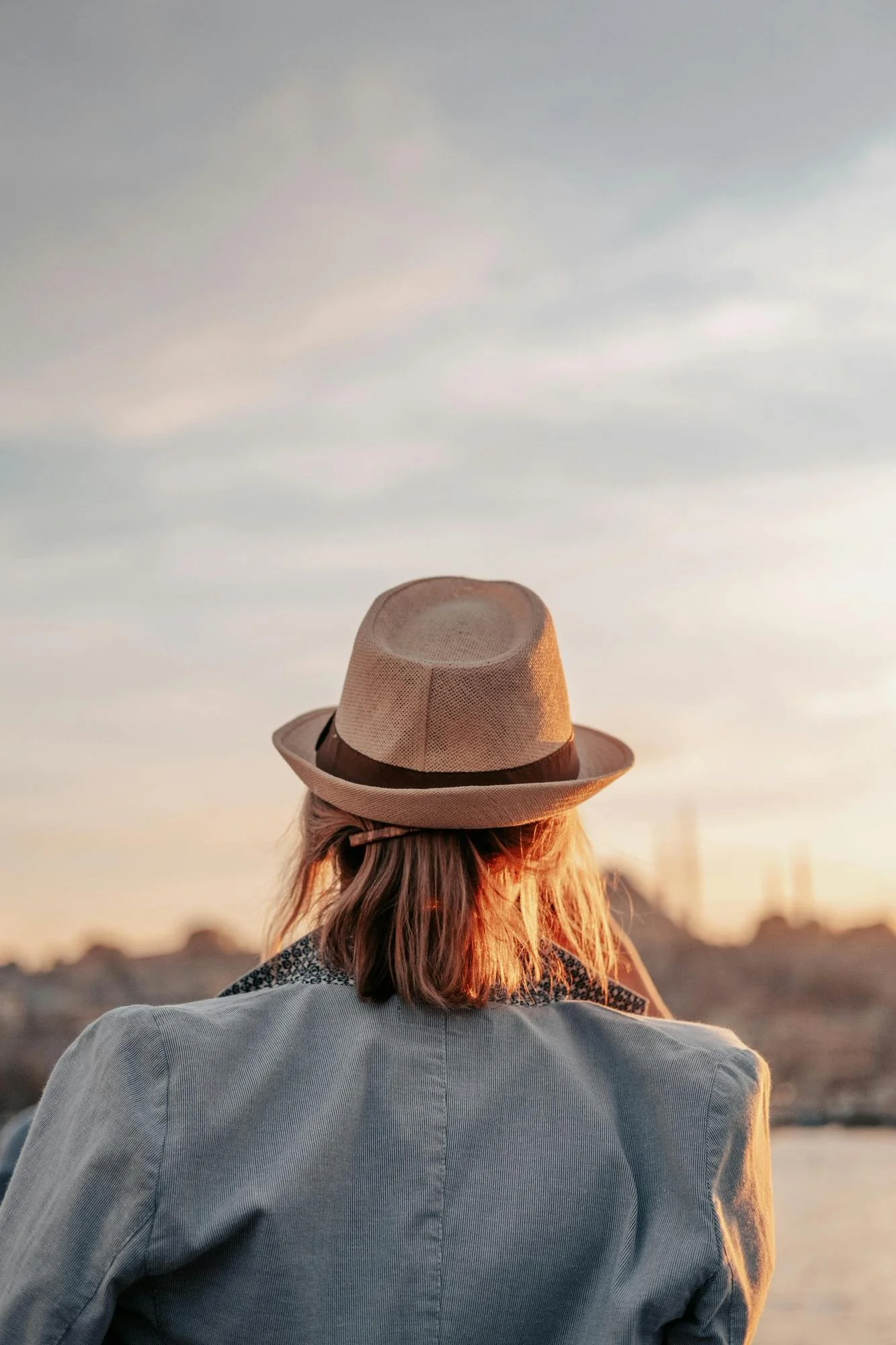 the back of blonde woman in hat facing the horizon during golden hour