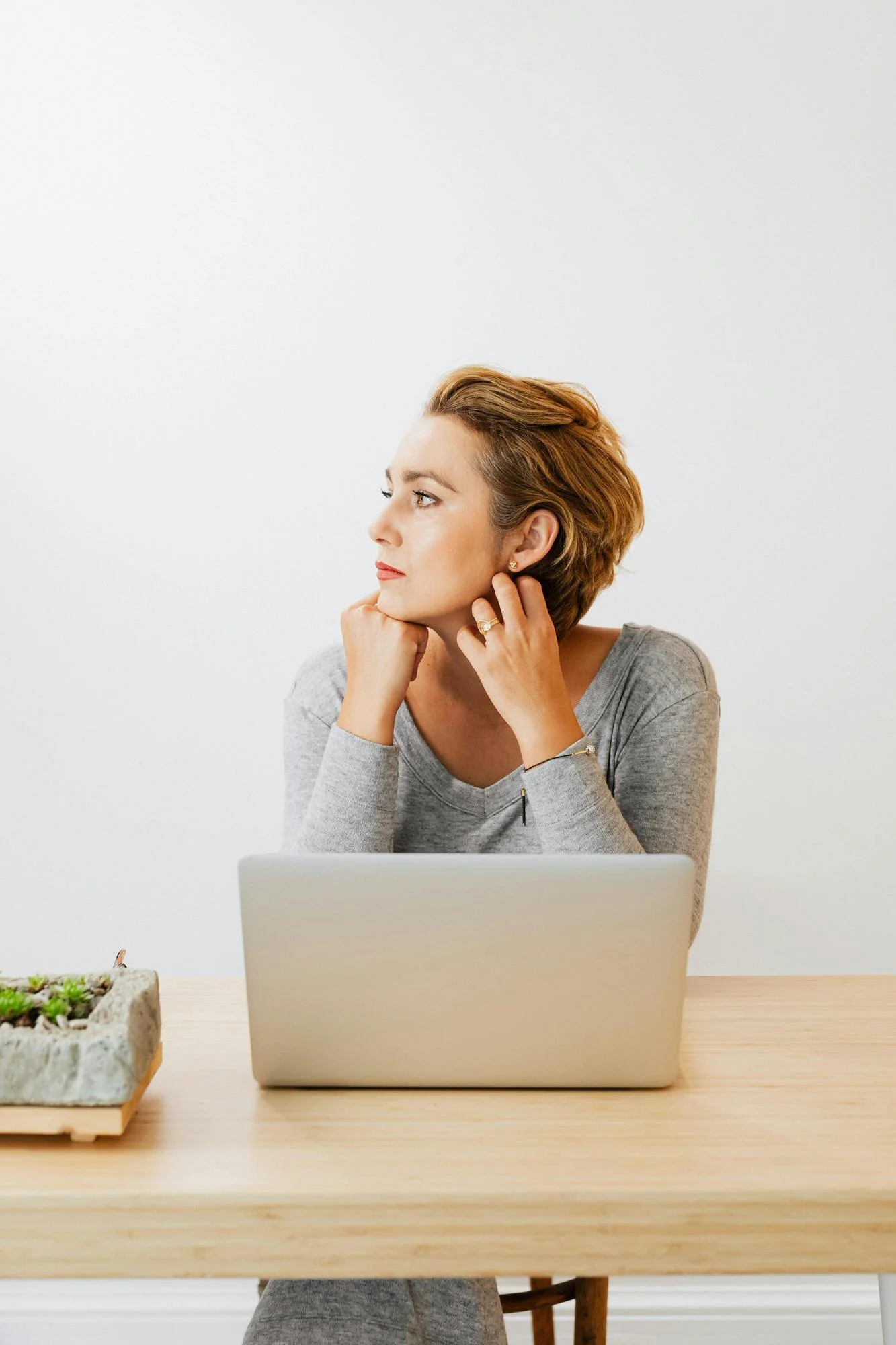 woman in gray shirt chin resting in hands looking off to the side with laptop on table in front of her