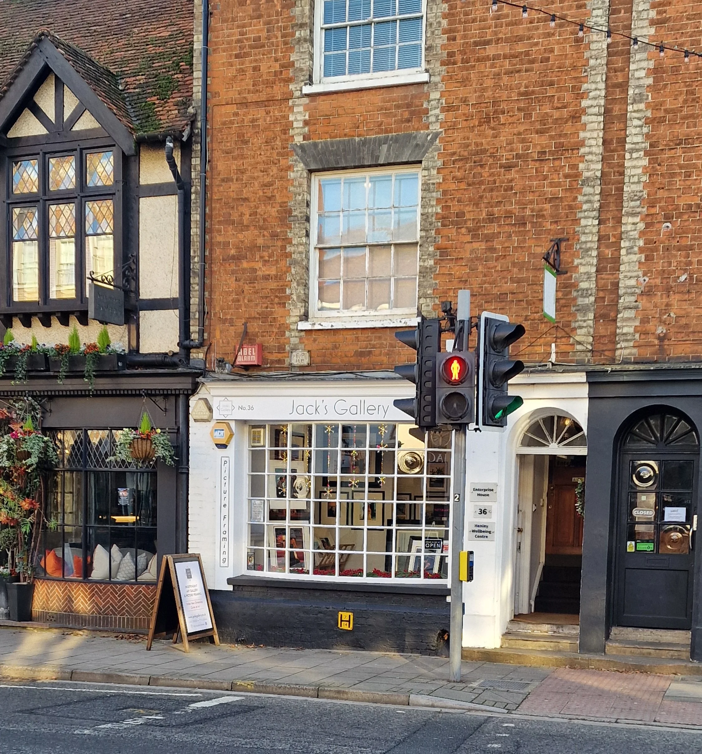 Street view showing a brick building with a gallery shop called "Jack's Gallery" at the ground level. There is a pedestrian traffic light showing red and a sidewalk cafe with potted plants and a menu board.