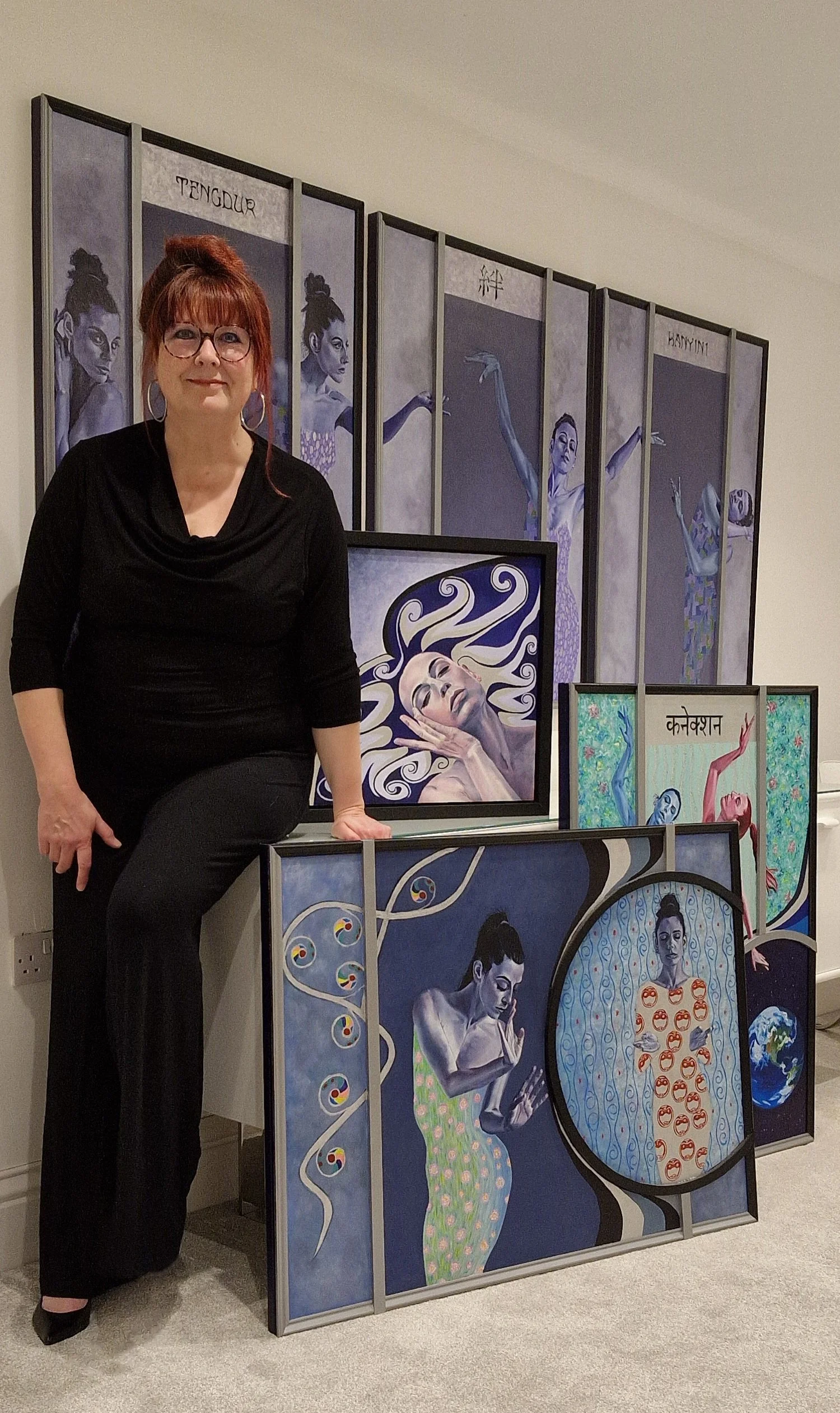A woman with red hair, glasses, and hoop earrings is standing next to a display of five colorful paintings featuring women in various artistic poses and styles, with Asian-inspired motifs and symbols, against a beige wall.