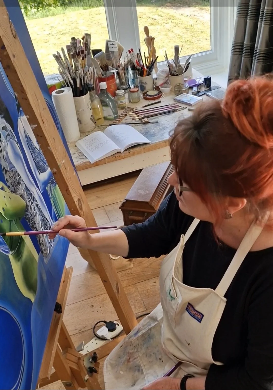 An artist with red hair and glasses working on a painting in a studio. The work area has various art supplies, brushes, and an open book. She wears a beige apron and is painting on a colorful canvas.