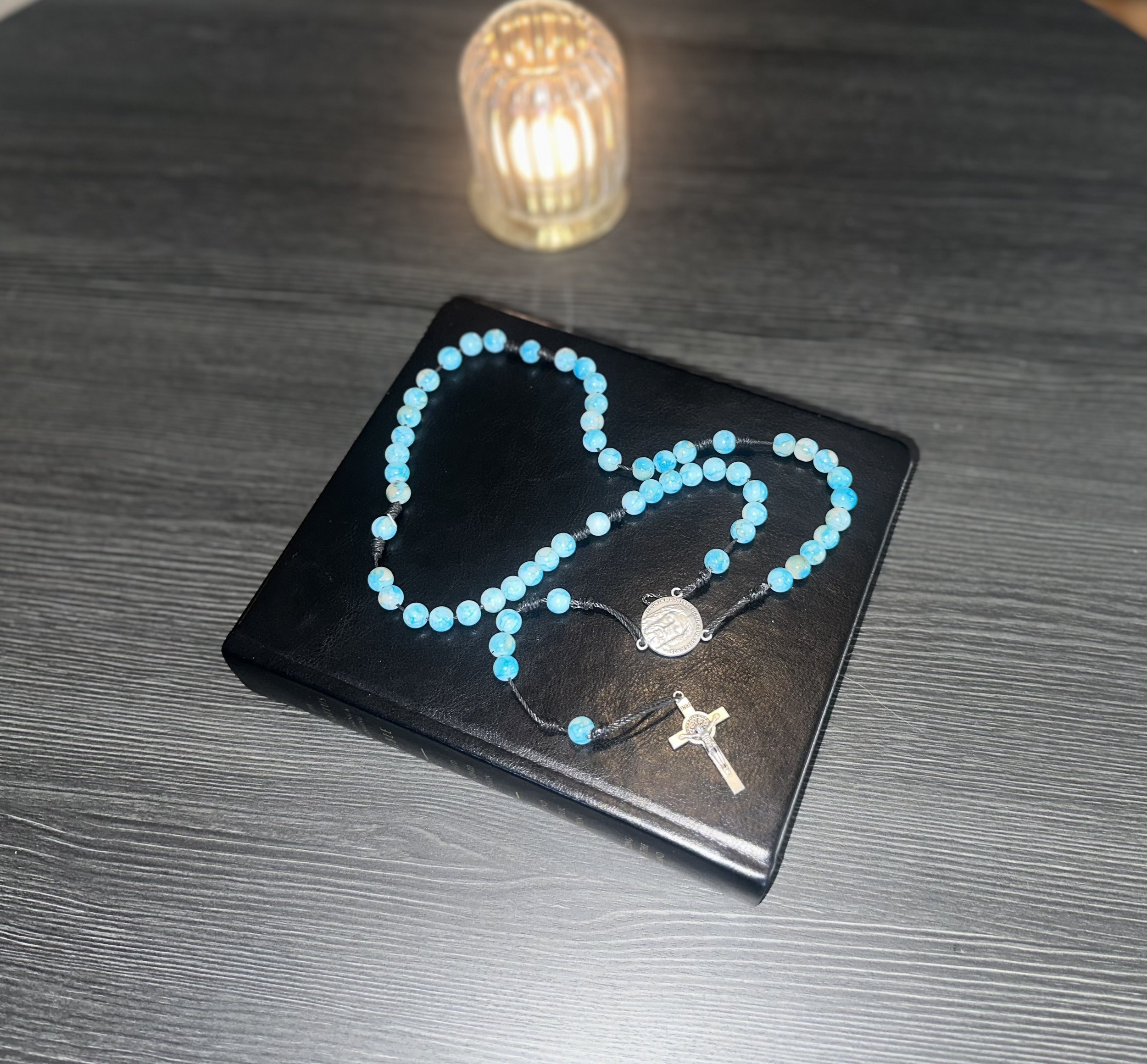 Vibrant turquoise rosary featuring a silver Jesus centerpiece and a St. Benedict medal, hand-knotted on a black cord and displayed on a black Ignatius Bible.
