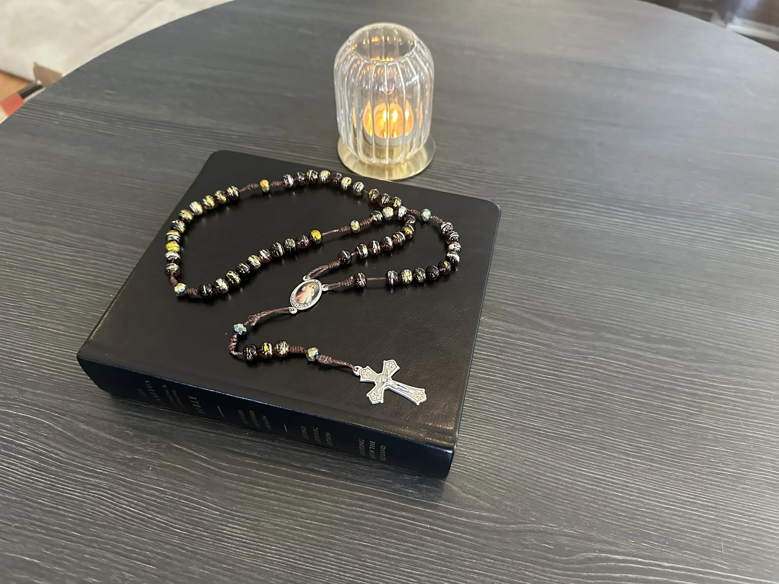 Hand-knotted Tiger's Eye rosary with a silver Resurrected Jesus centerpiece, resting on a black leather Bible next to a glowing candle.