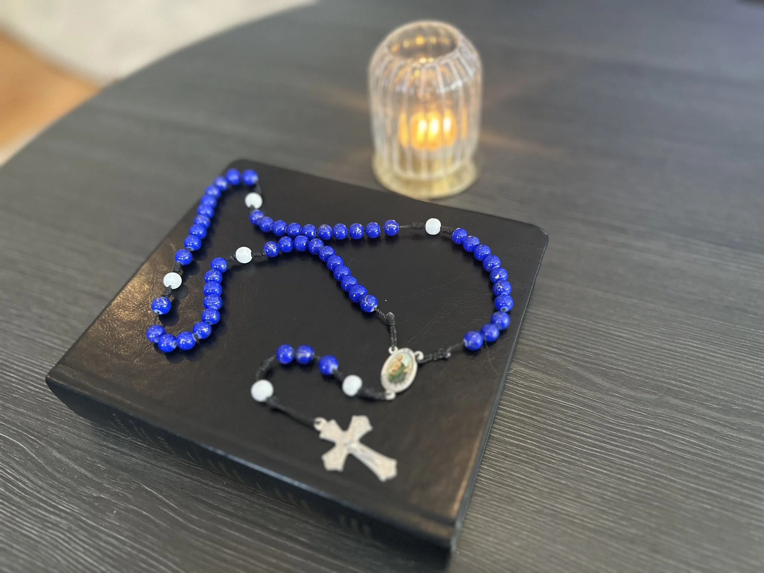 Hand-knotted rosary with deep blue and white beads on a black cord, featuring a colorful St. Jude Thaddeus centerpiece, resting on a black leather Bible.