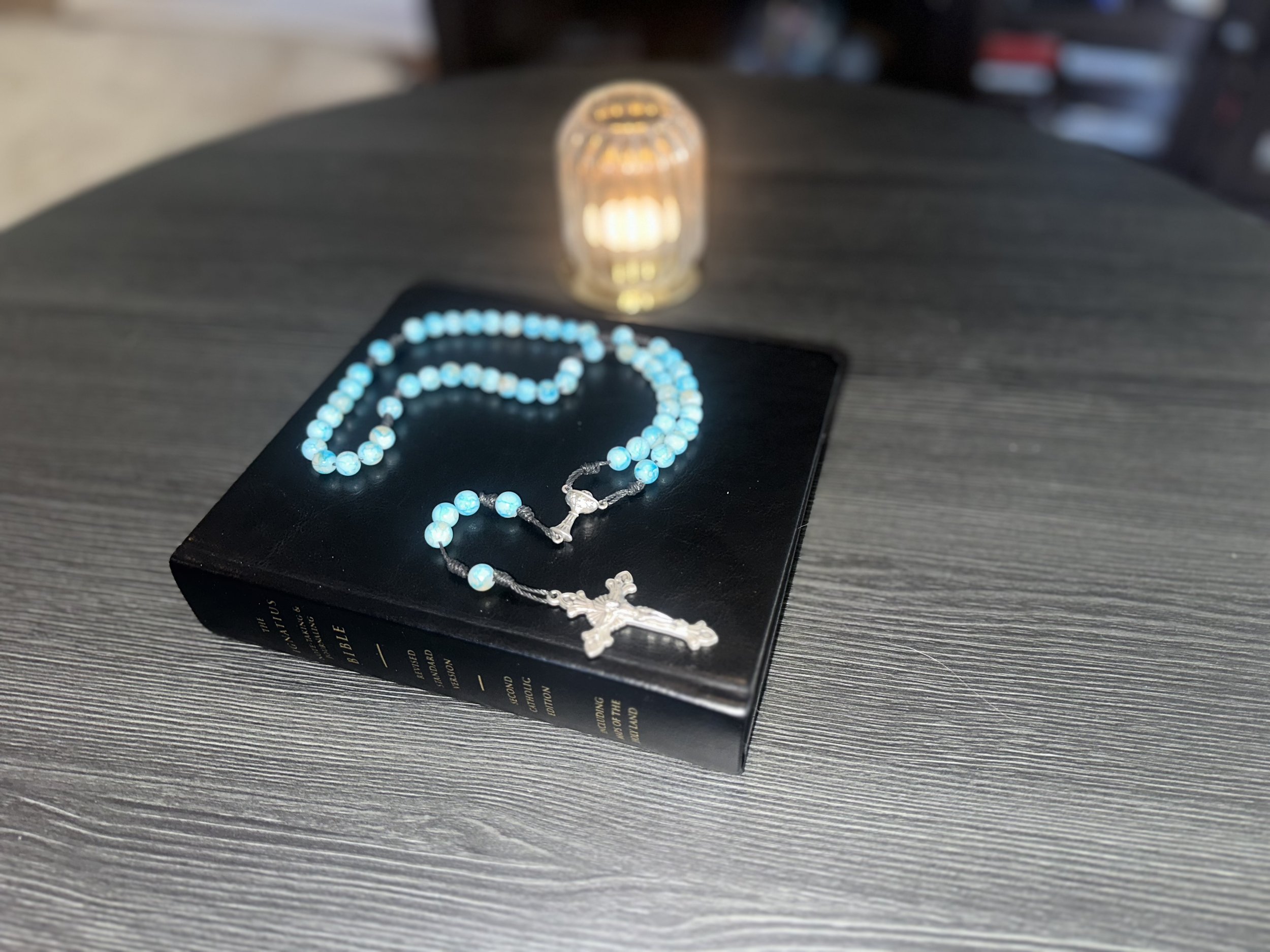 Hand-knotted turquoise rosary with mottled blue-green beads and a silver Chalice centerpiece, resting on a black leather Bible next to a glowing candle.
