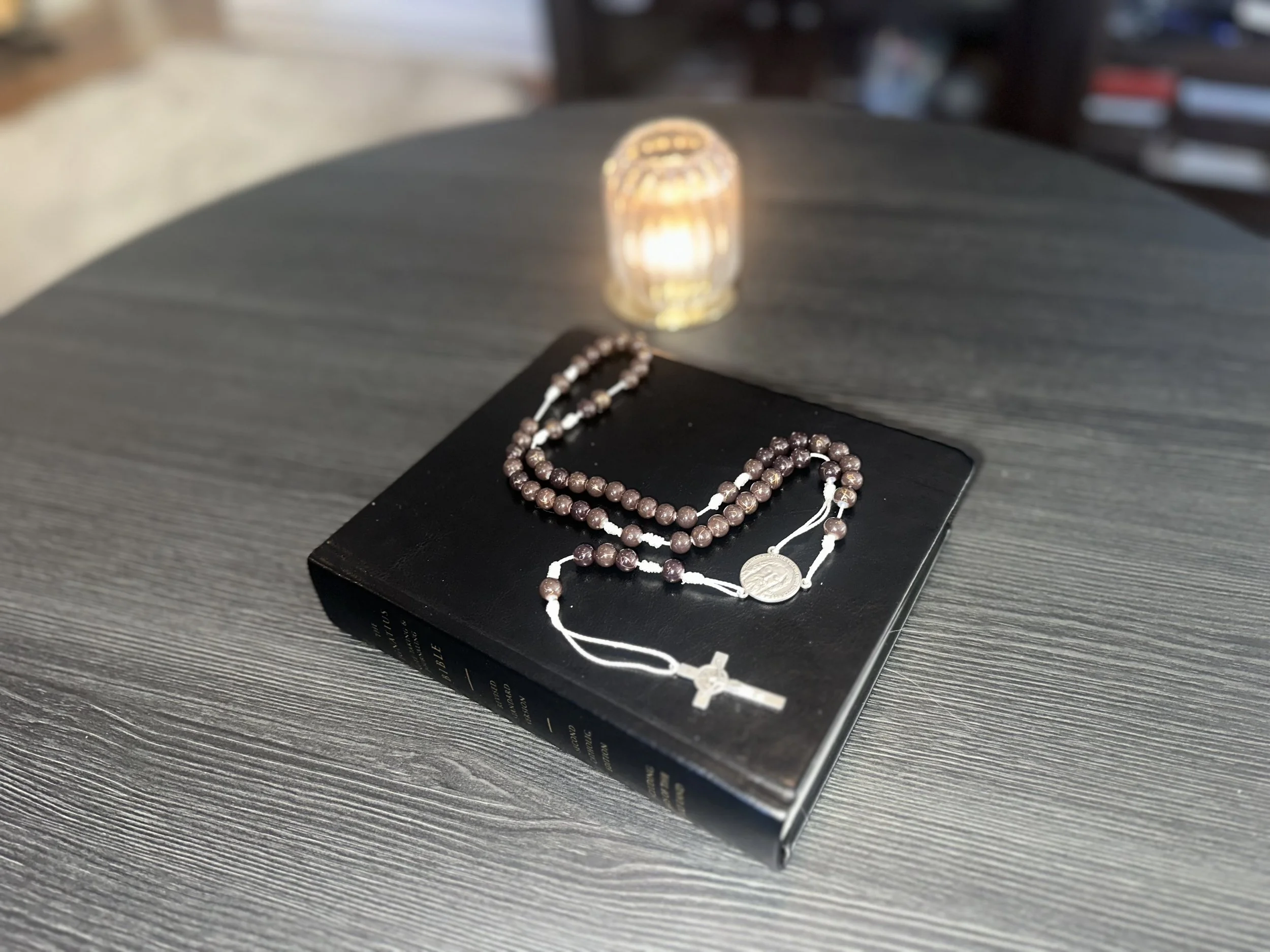 Hand-knotted rosary with dark brown beads and a white cord, featuring a Jesus centerpiece, resting on a black Bible.