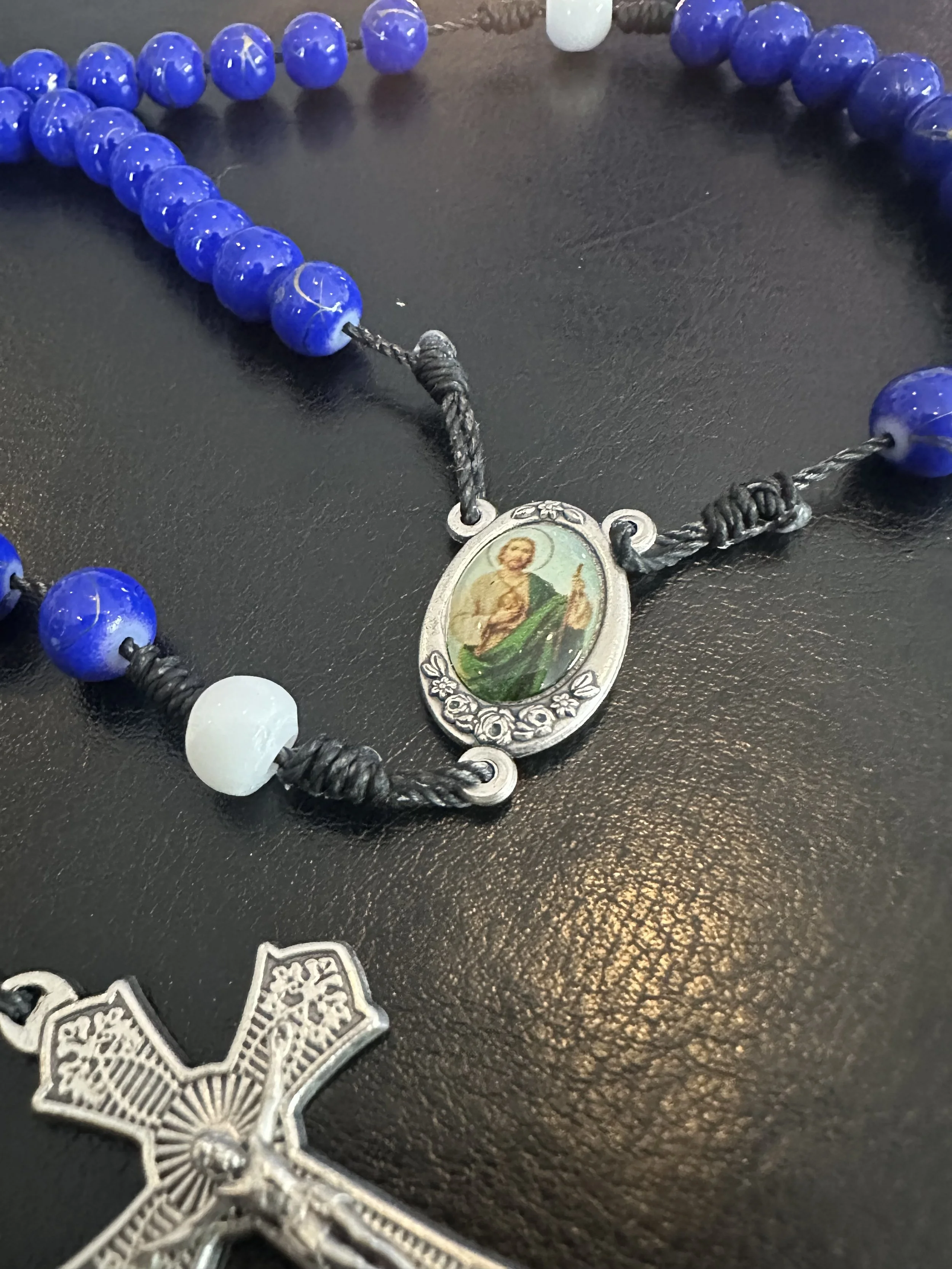 Detailed close-up of a silver St. Jude Thaddeus centerpiece with green and gold enamel, shown on a hand-knotted rosary with vibrant blue and white beads.