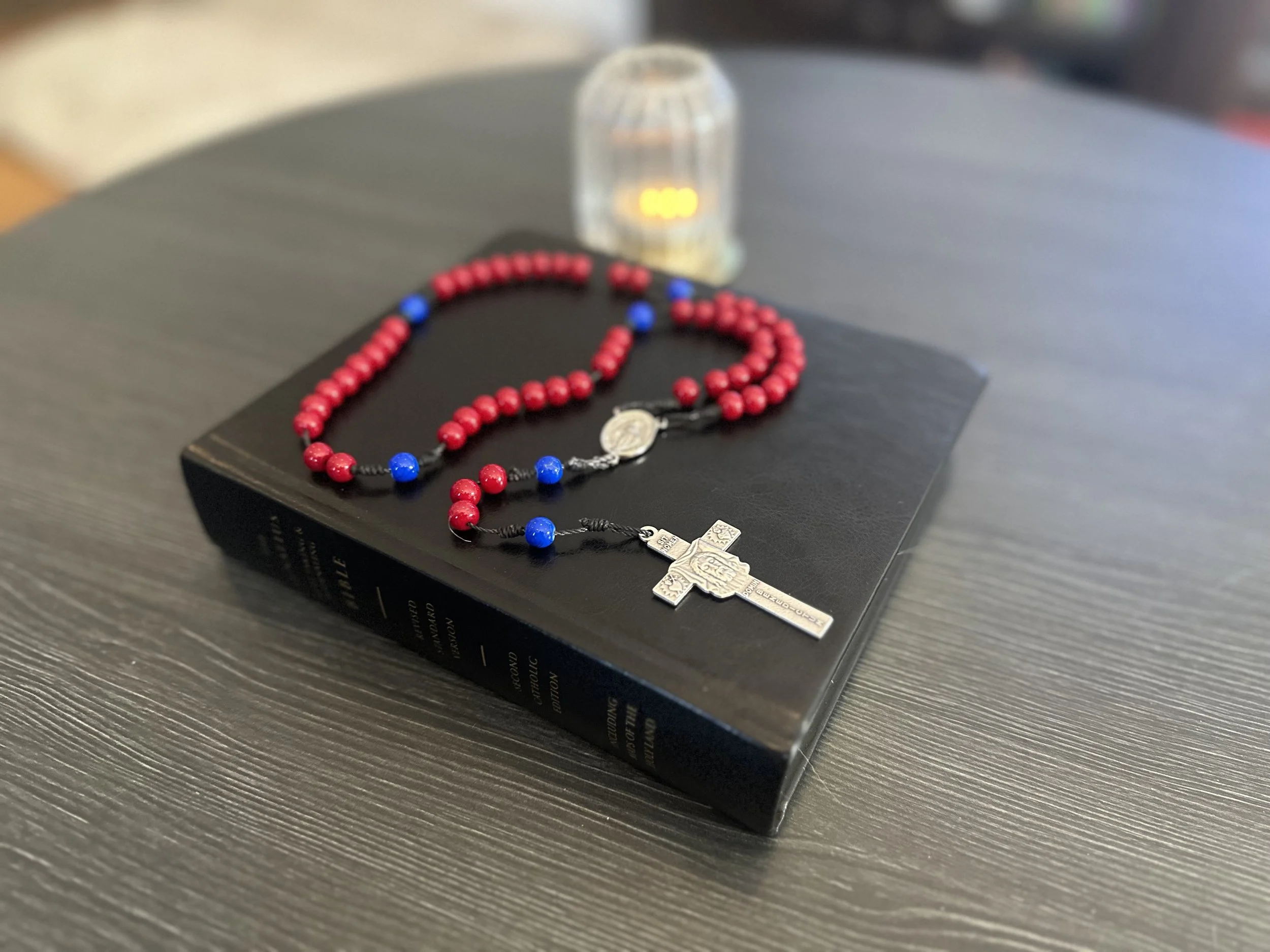 Hand-knotted rosary with red beads and blue accent beads on a black cord, featuring a silver Miraculous Medal centerpiece and a detailed St. Benedict crucifix, displayed on a black Ignatius Bible.