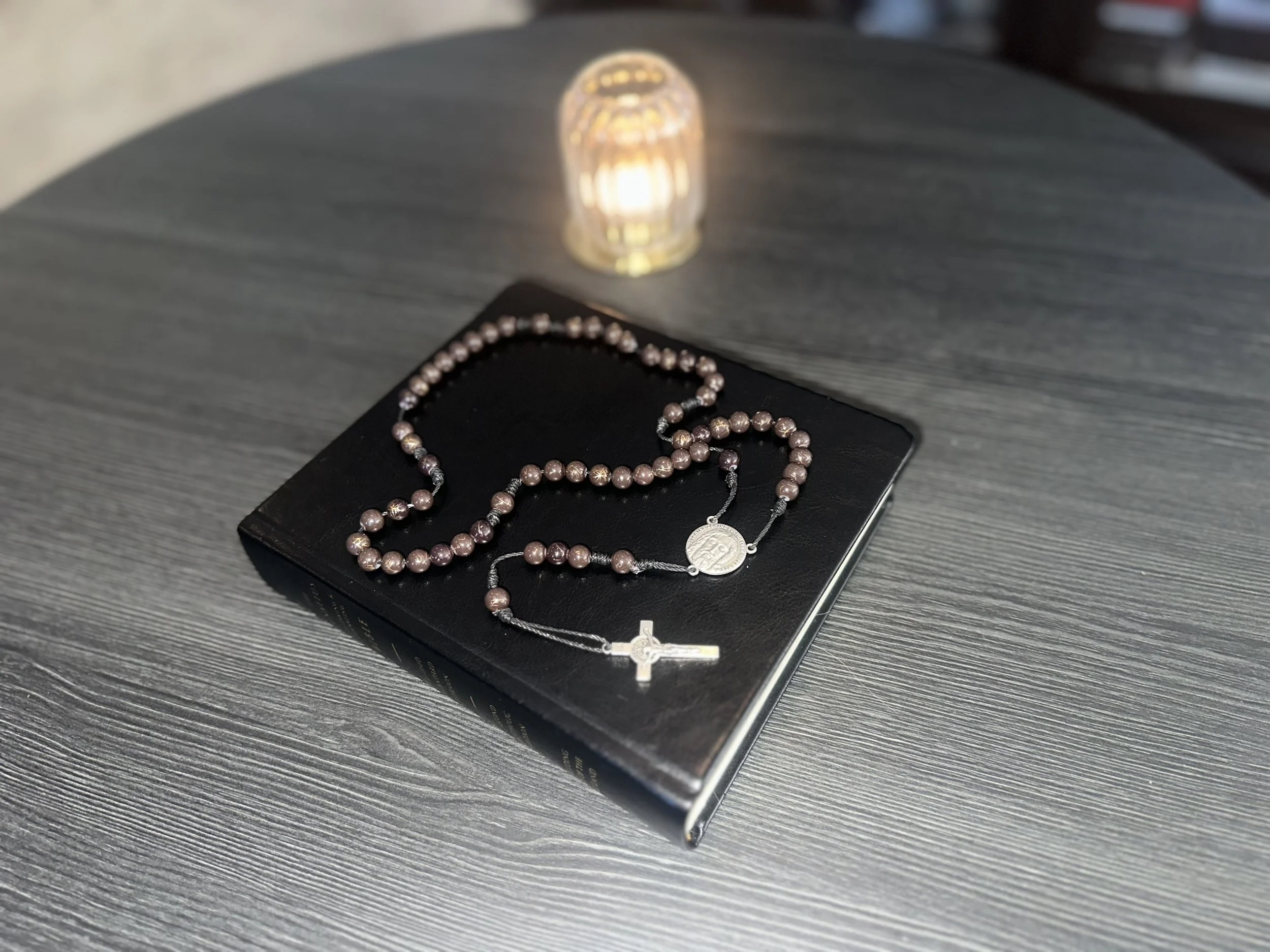 Hand-knotted rosary with dark brown beads and a sturdy black cord, featuring a Jesus centerpiece, displayed next to a lit candle.