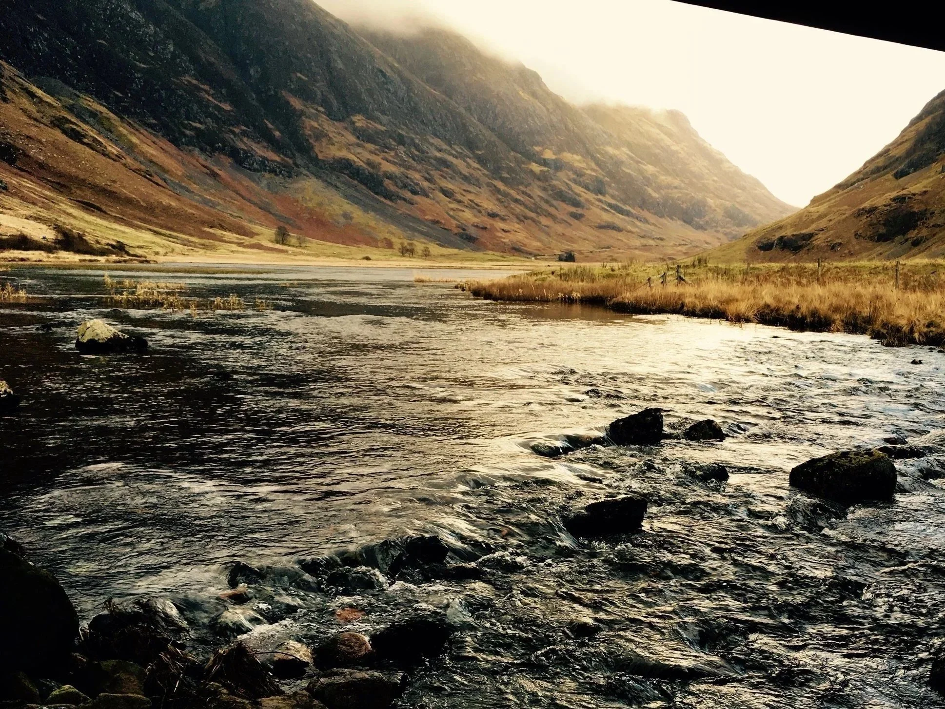 view of Glencoe valley in the Scottish Highlands, with the Three Sisters mountains rising above the winding A82 road under dramatic clouds.
