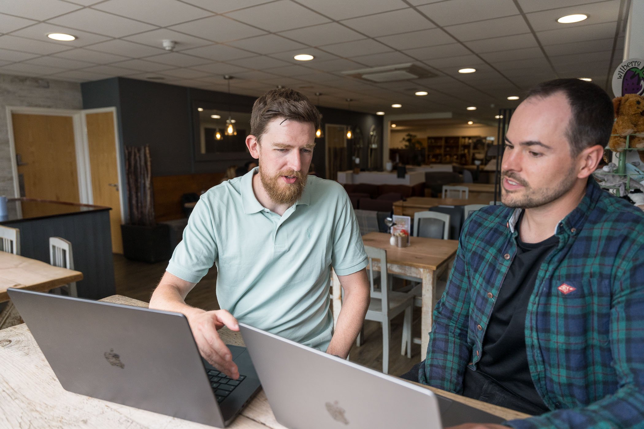 Two men sitting at a table in a café working on their laptops, engaged in conversation.