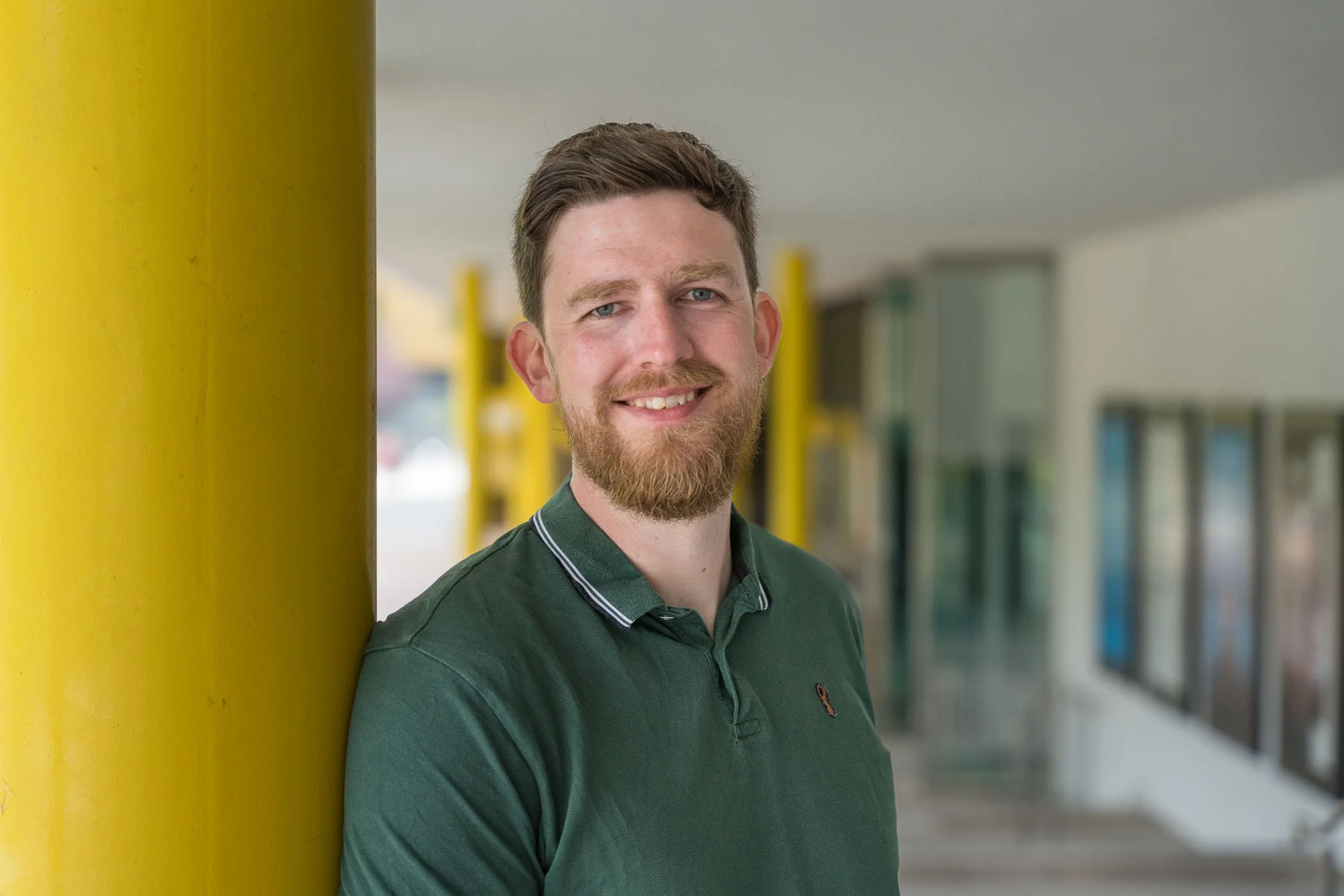A smiling man with a beard and short brown hair leaning against a yellow pole in an outdoor setting with a blurred background.