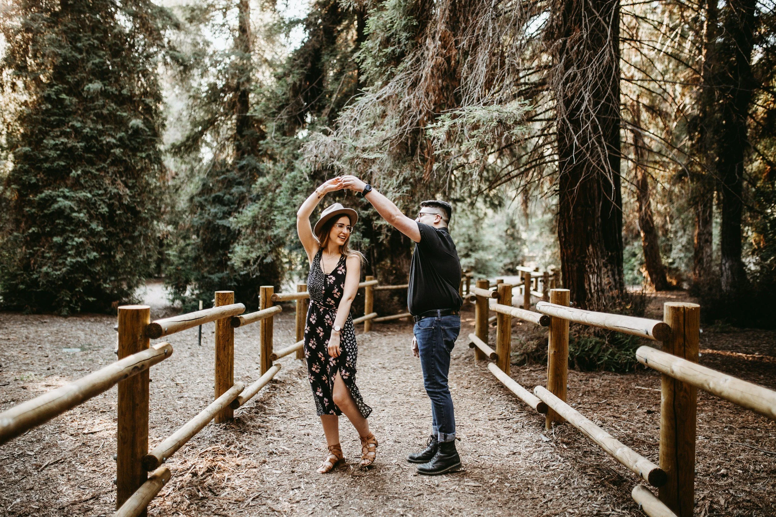 A couple dancing together on a dirt path in a forest, surrounded by tall trees and greenery, with wooden fences on either side.