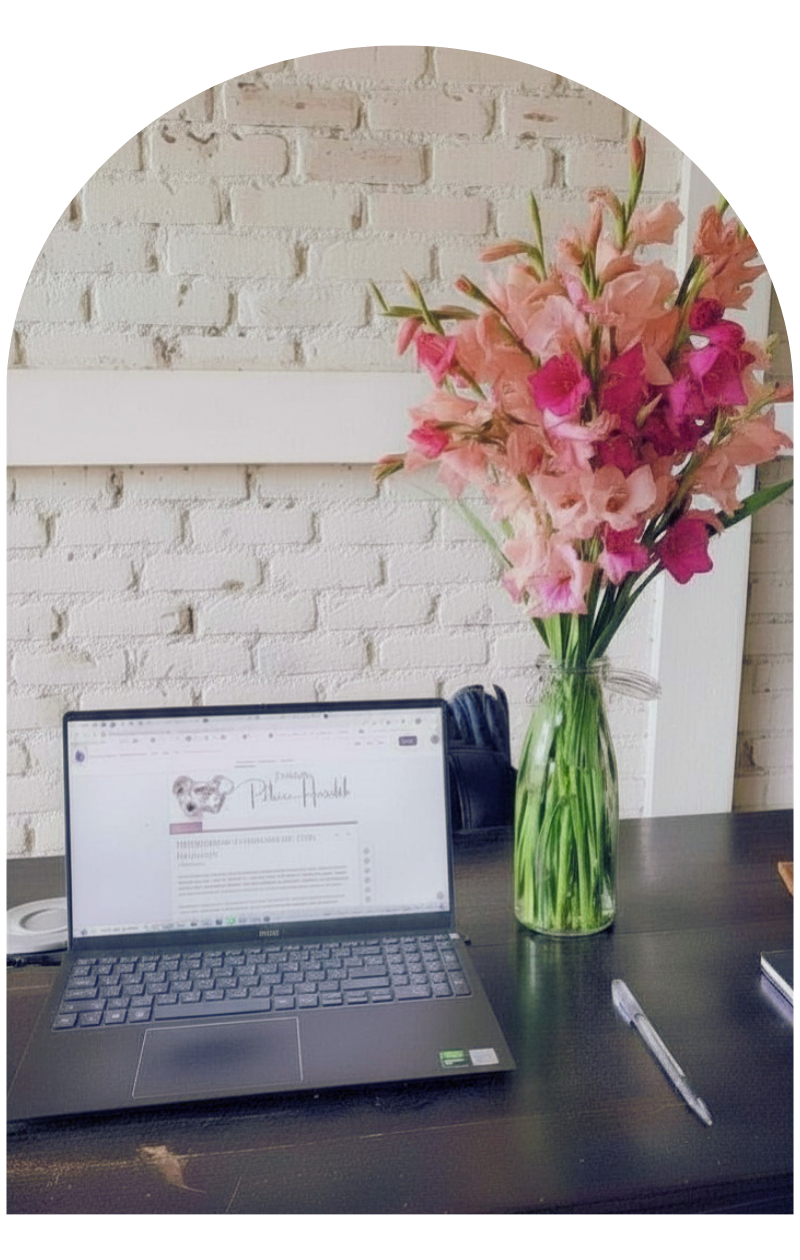 A computer laptop on a dark desk with a vase of pink and peach flowers beside it, set against a white brick wall.