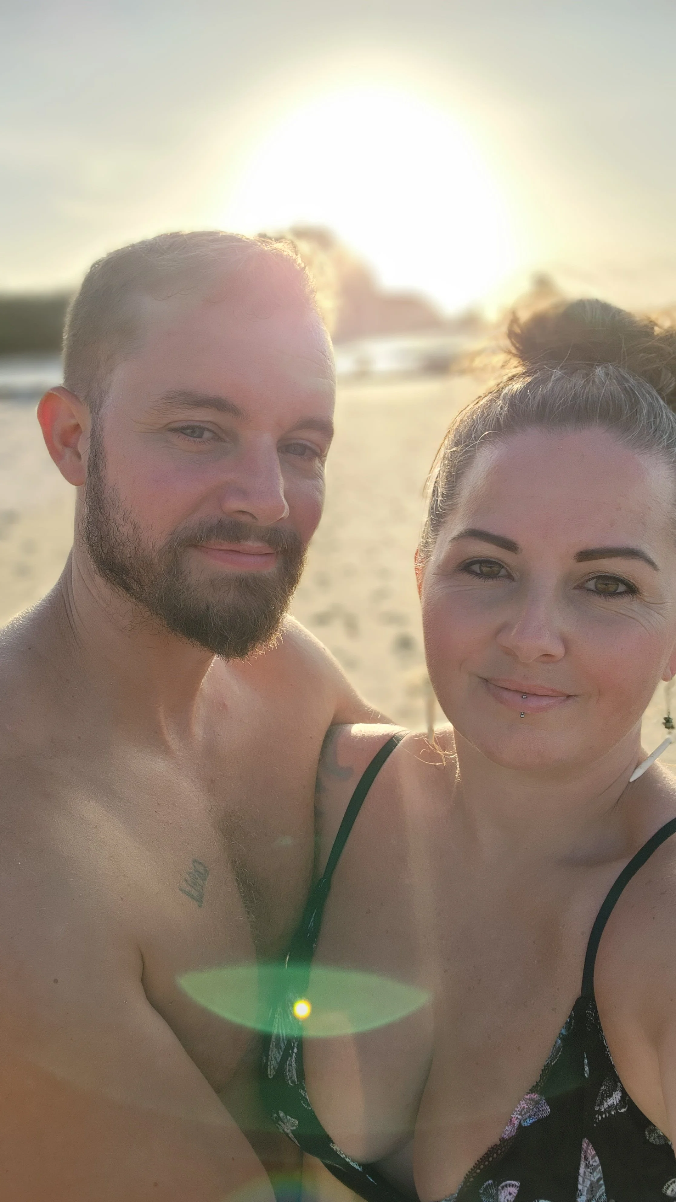 A man and woman taking a selfie on a sandy beach during sunset, with the sun setting behind them and a lens flare visible in the foreground.