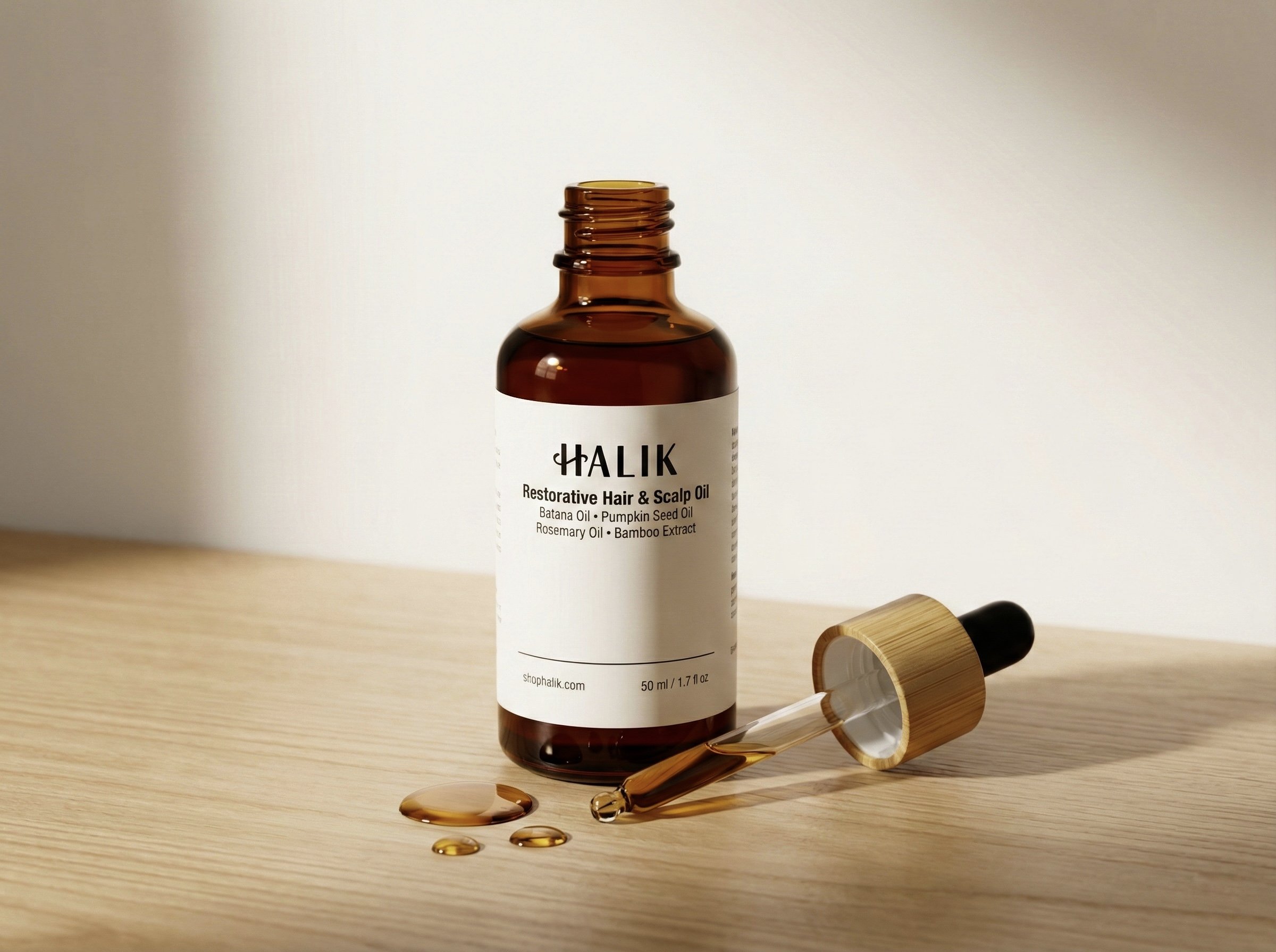A brown glass bottle labeled 'HALIK Restorative Hair & Scalp Oil' with a dropper beside it, containing some oil, and oil droplets on a beige surface