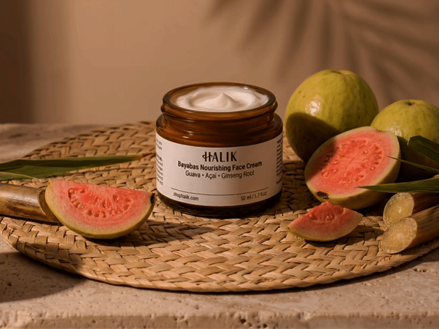 An amber jar of luxury botanical face cream with a minimalist white label on top of a woven mat and surrounded by guavas and sugarcane pieces