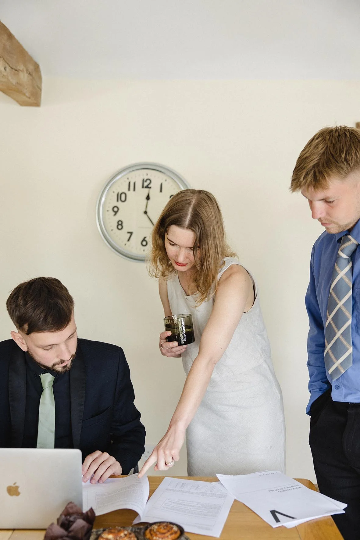 A woman in a sleeveless white dress points at some documents on a table while talking to two men in professional attire, one seated with a laptop and the other standing. The setting appears to be an office during a work discussion, with a large clock showing 11:55 on the wall.