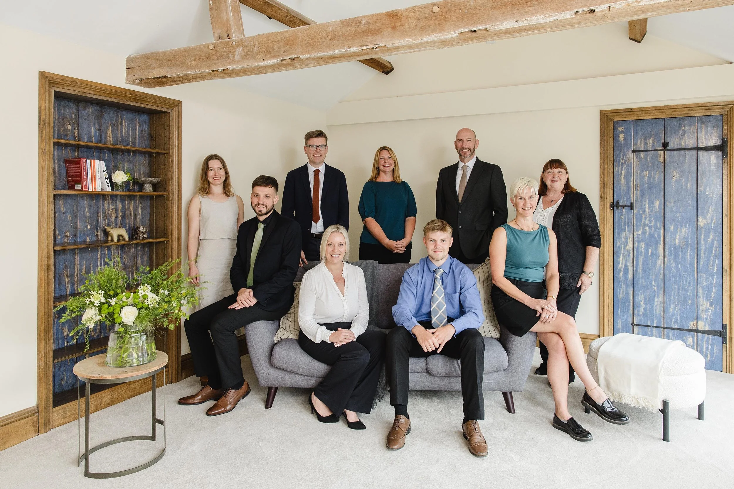 Group of ten professionally dressed people posing in a cozy living room with wooden accents, a gray sofa, a bookshelf, and decorative plants.