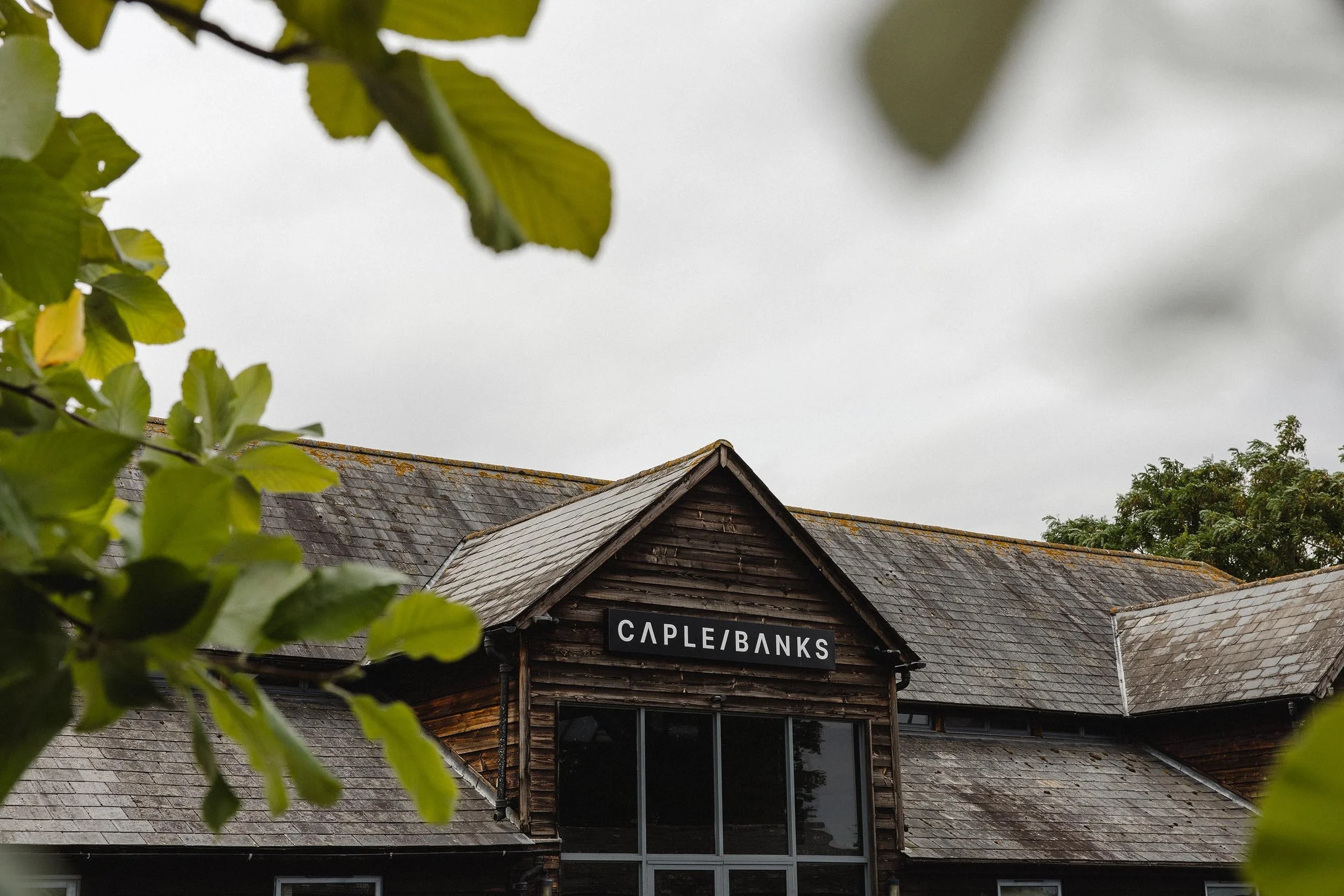 The image shows a rustic wooden building with a sign that says 'Cape Banks' on the front. The building has a peaked roof with shingles and is partially obscured by green leaves in the foreground. The sky above is cloudy.