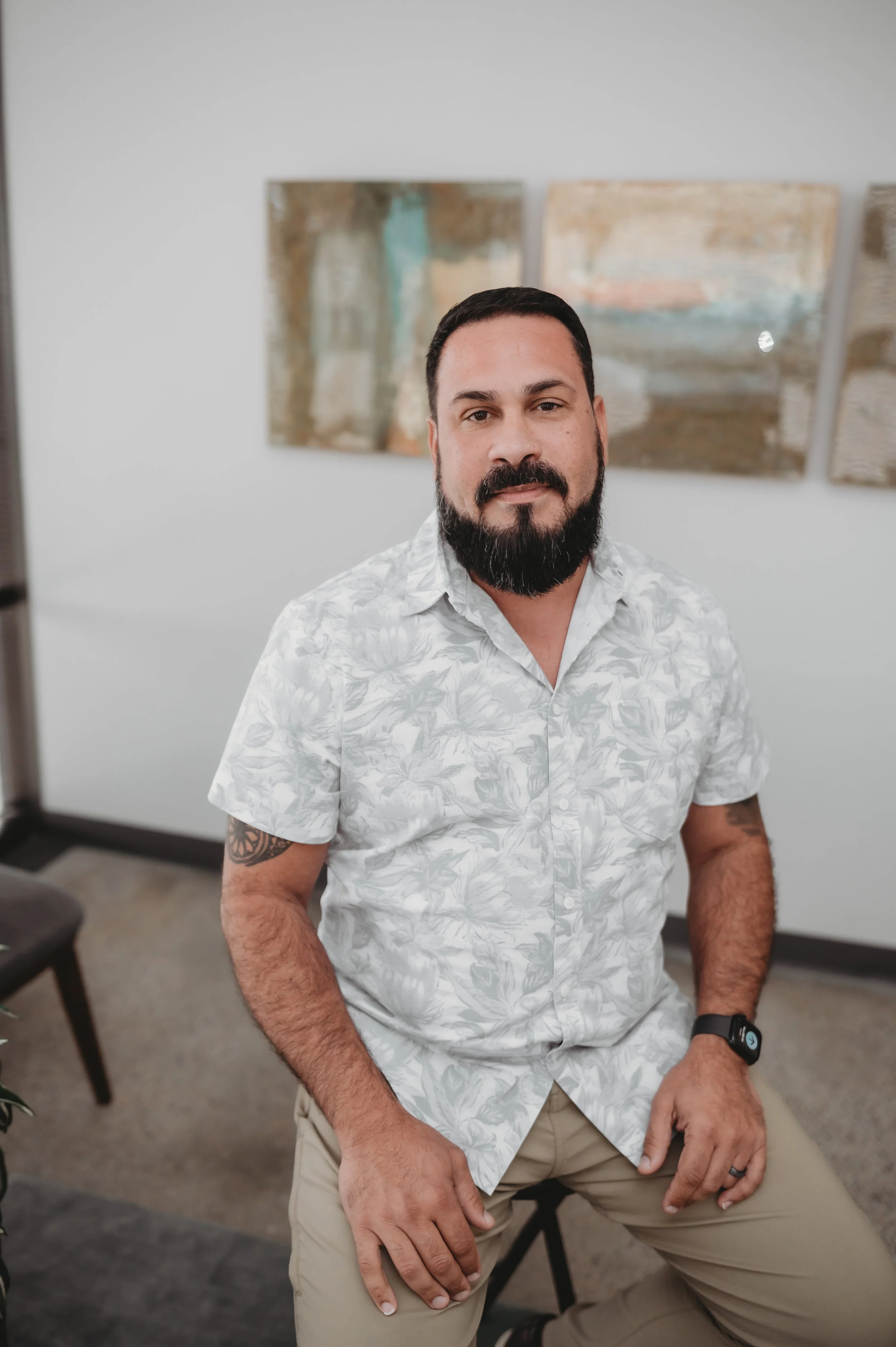 A man with a beard and dark hair, wearing a white floral short-sleeve shirt, sitting in a room with abstract paintings on the wall behind him.