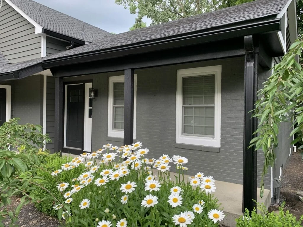 A house with gray brick exterior, white trim, black front door, two window panes, small porch with black supports, surrounded by green shrubbery and white daisies in the garden.
