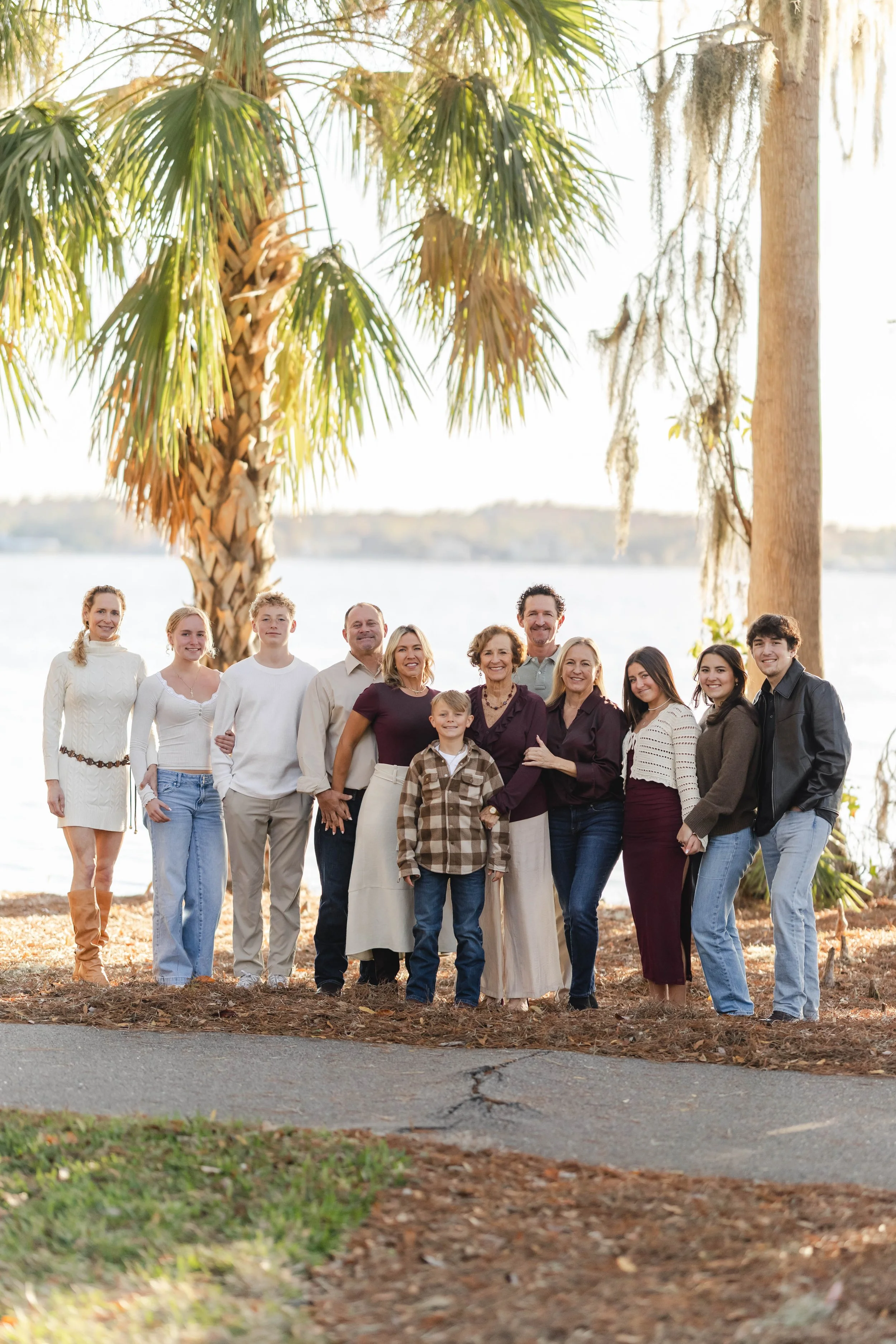 A large family standing outdoors in front of a lake, surrounded by trees, with a mix of adults and children dressed casually for fall or mild weather.