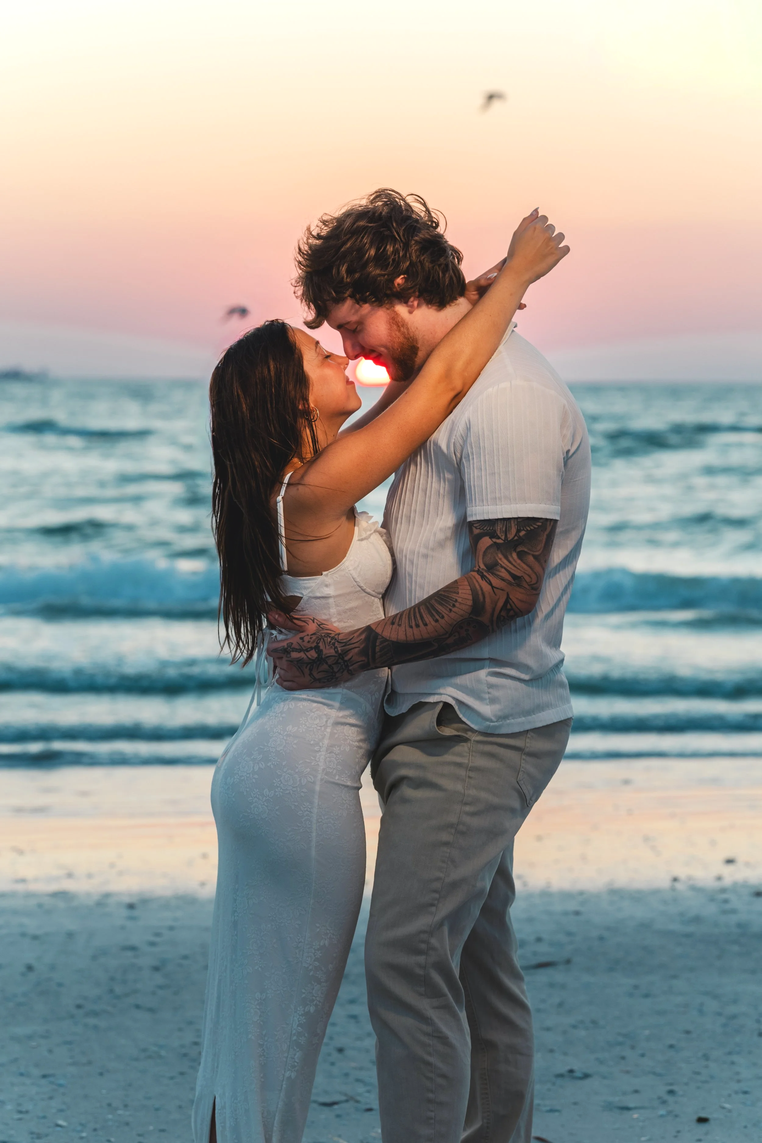 A couple embraces on the beach at sunset, the woman in a white dress and the man with tattoos, their foreheads touching, with the ocean and setting sun in the background.