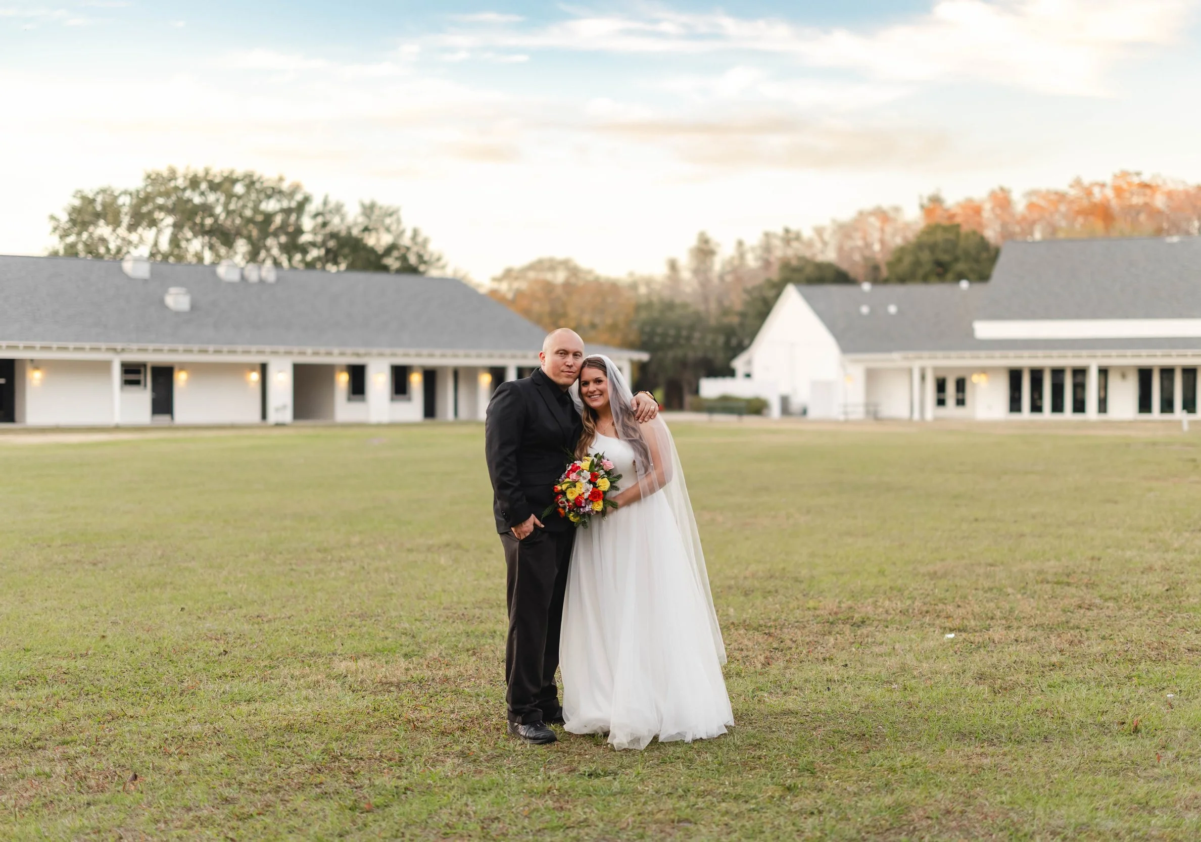 A newlywed couple standing outdoors on a grassy field, with the groom in a black suit and the bride in a white wedding dress holding a bouquet of flowers, and a row of buildings and trees in the background during sunset.
