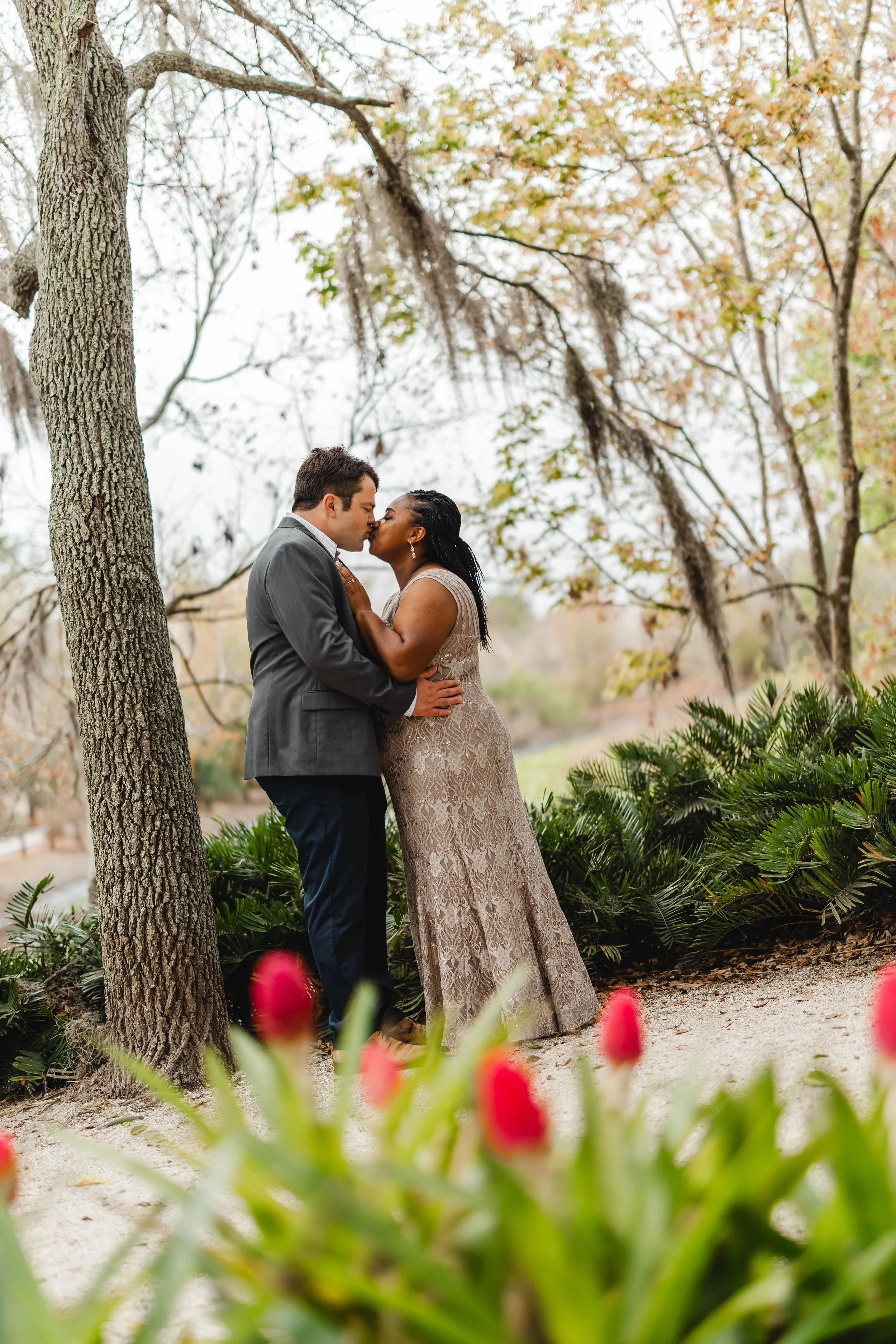 A couple sharing a kiss outdoors among trees and greenery, with a woman in a lace dress and a man in a gray suit, in a natural setting with blurred pink flowers in the foreground.
