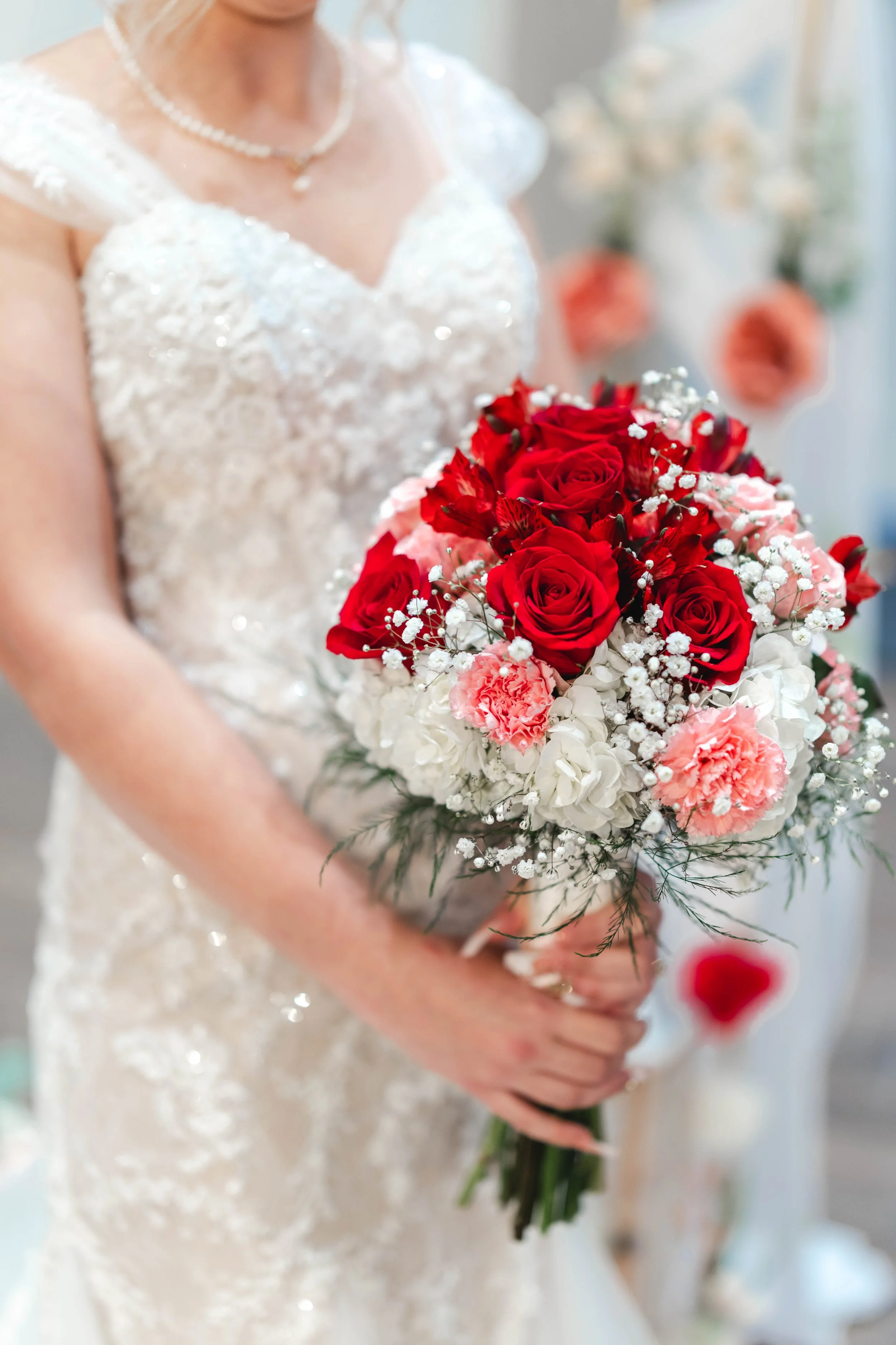 Bride in a white lace wedding dress holding a bouquet of red, pink, and white flowers.
