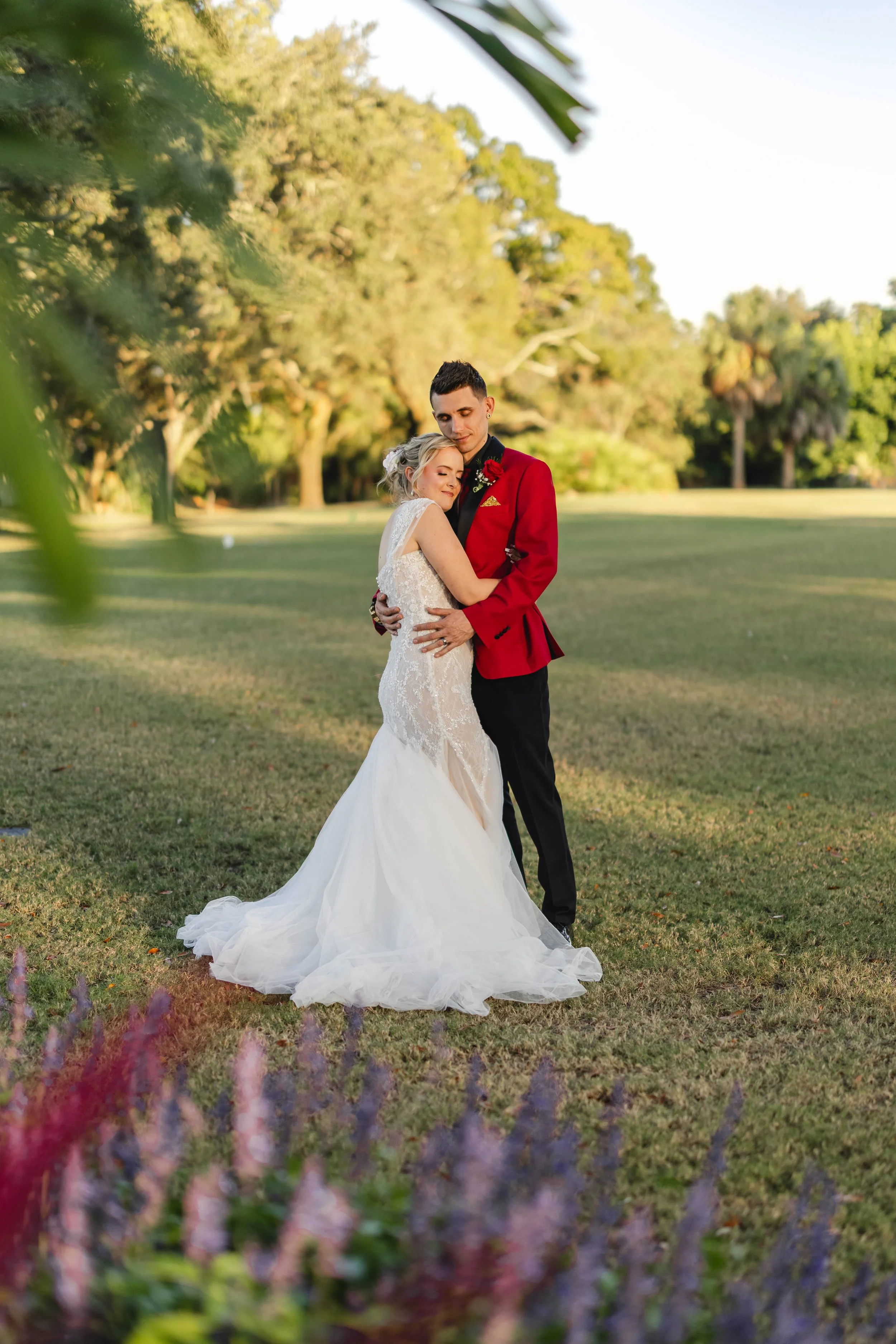 A bride and groom embrace on a grassy field during sunset, with trees in the background and colorful flowers in the foreground.