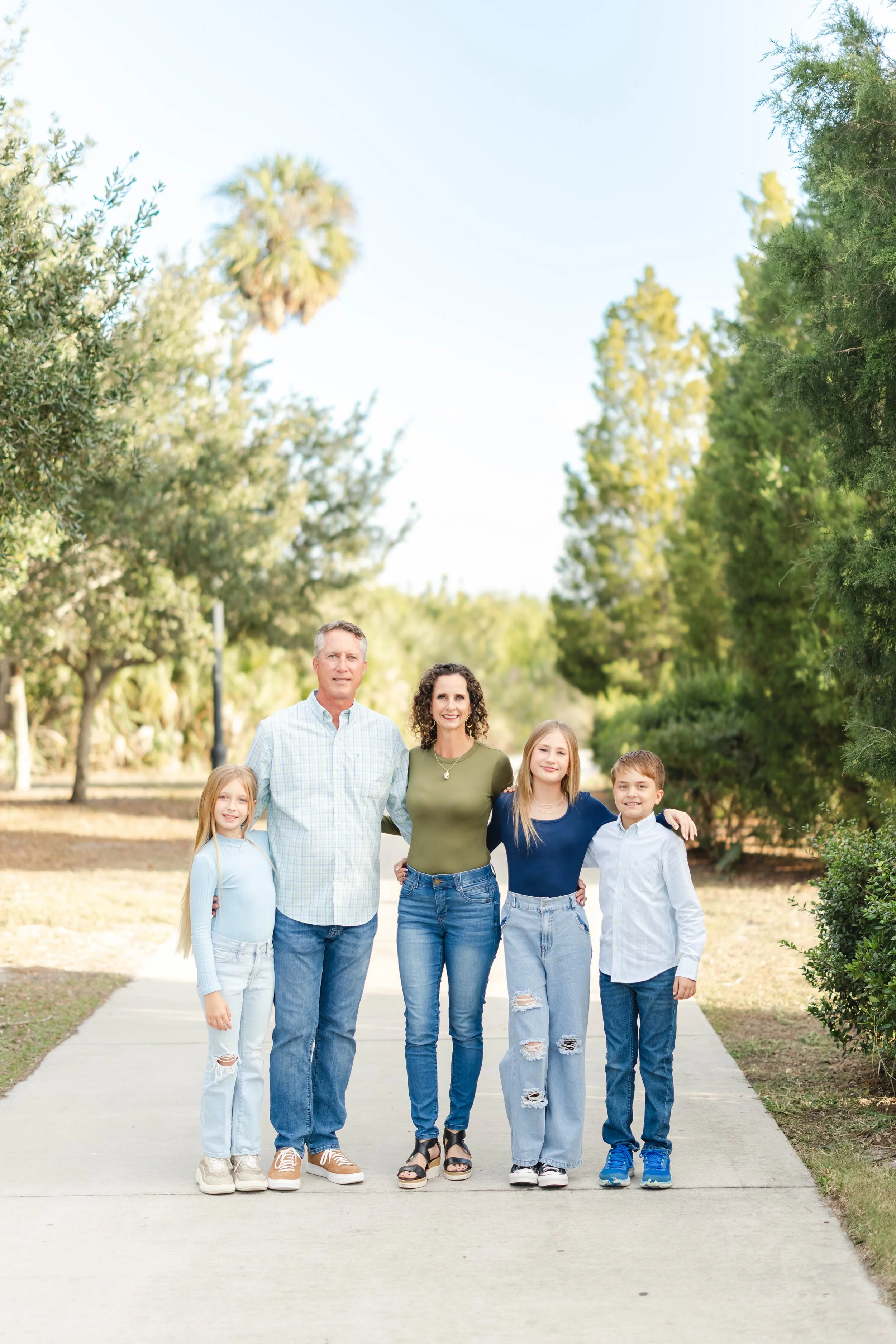 Family of five standing on a sidewalk in a green park, smiling, with trees and clear sky in the background.