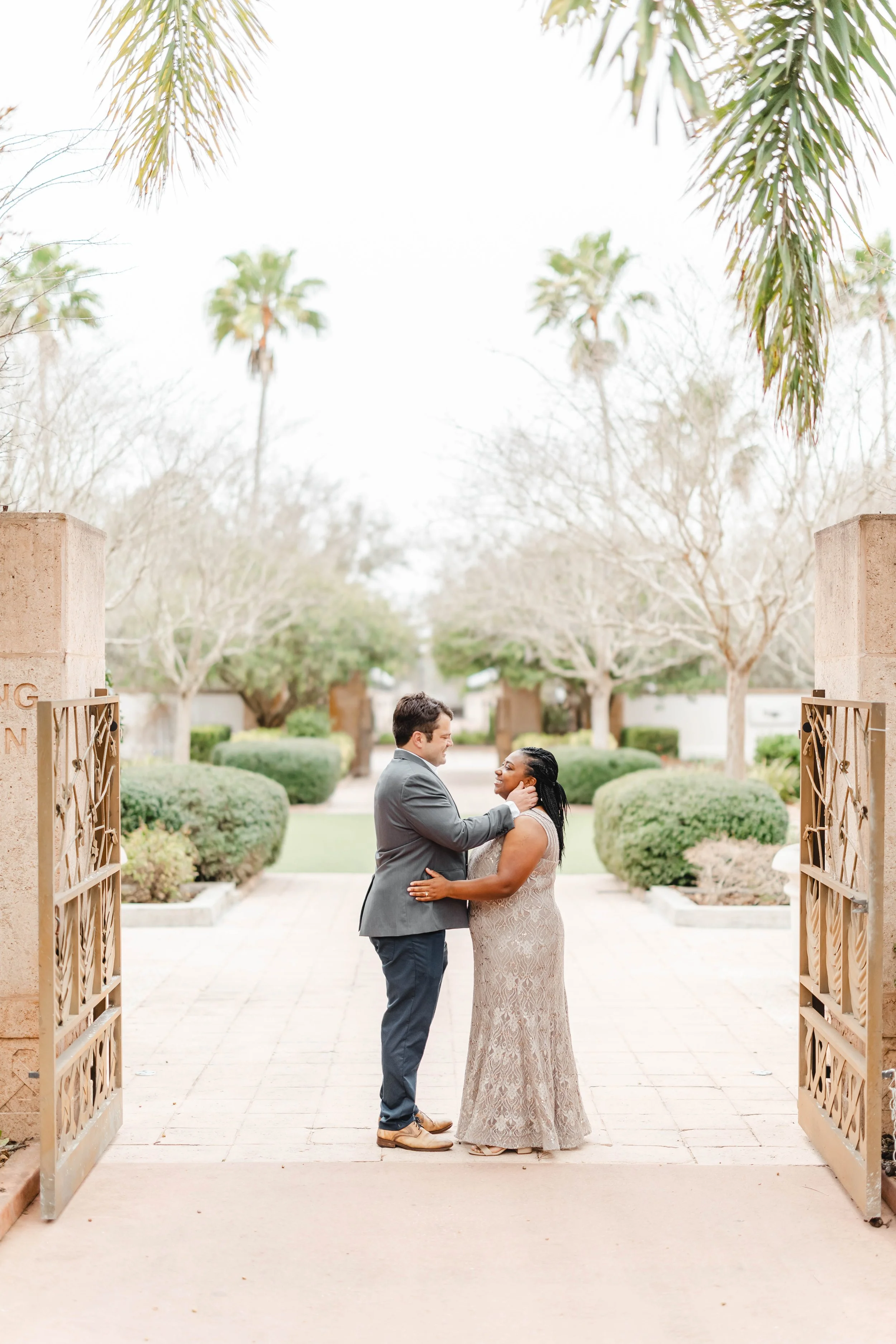 A couple stands facing each other in a romantic pose at an outdoor location, surrounded by landscaped bushes, trees, and palm trees in the background. The man is wearing a gray suit and the woman is dressed in a long, intricate lace gown.