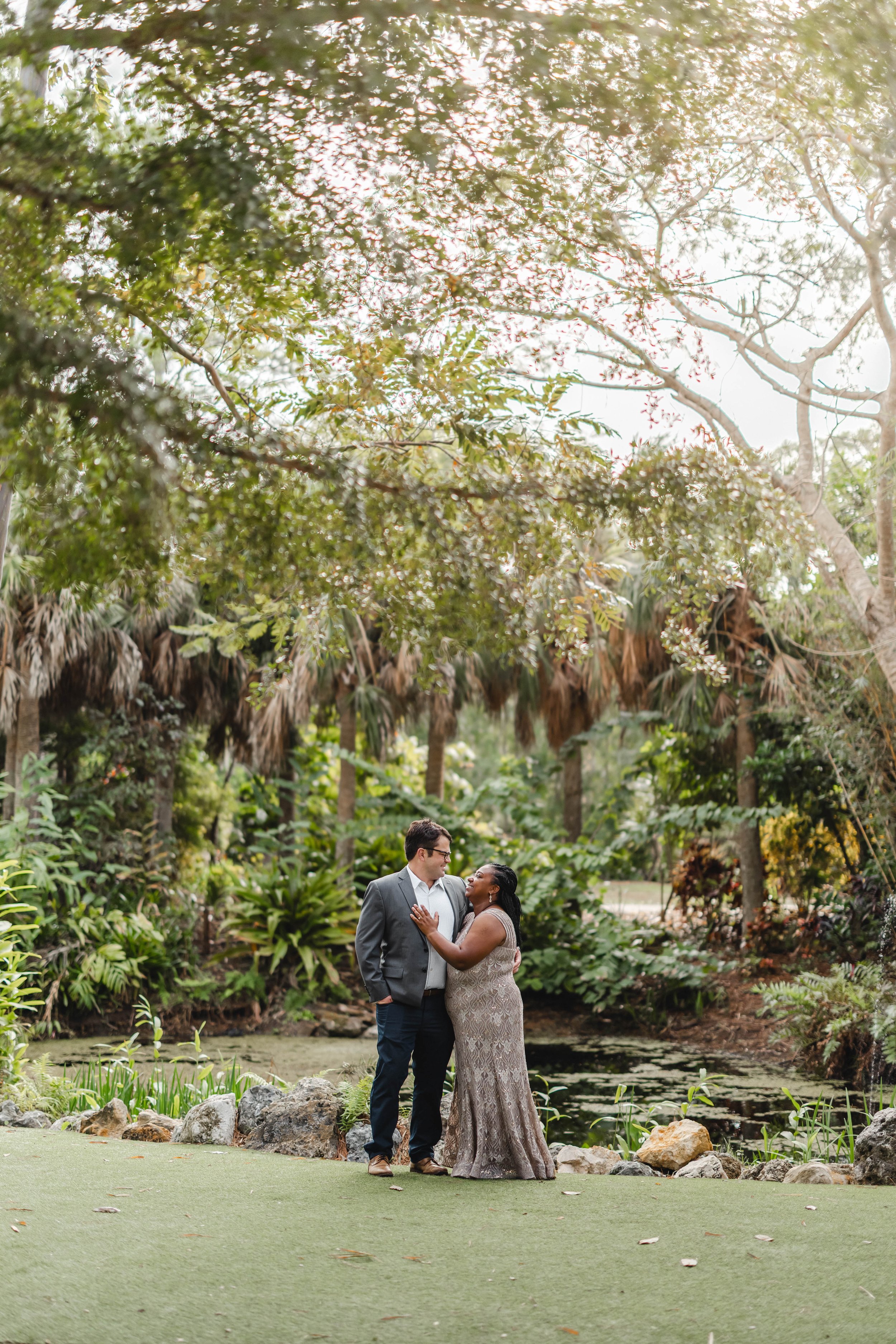 A couple standing close together outdoors in a lush green environment, with a pond and dense trees in the background, during daytime.