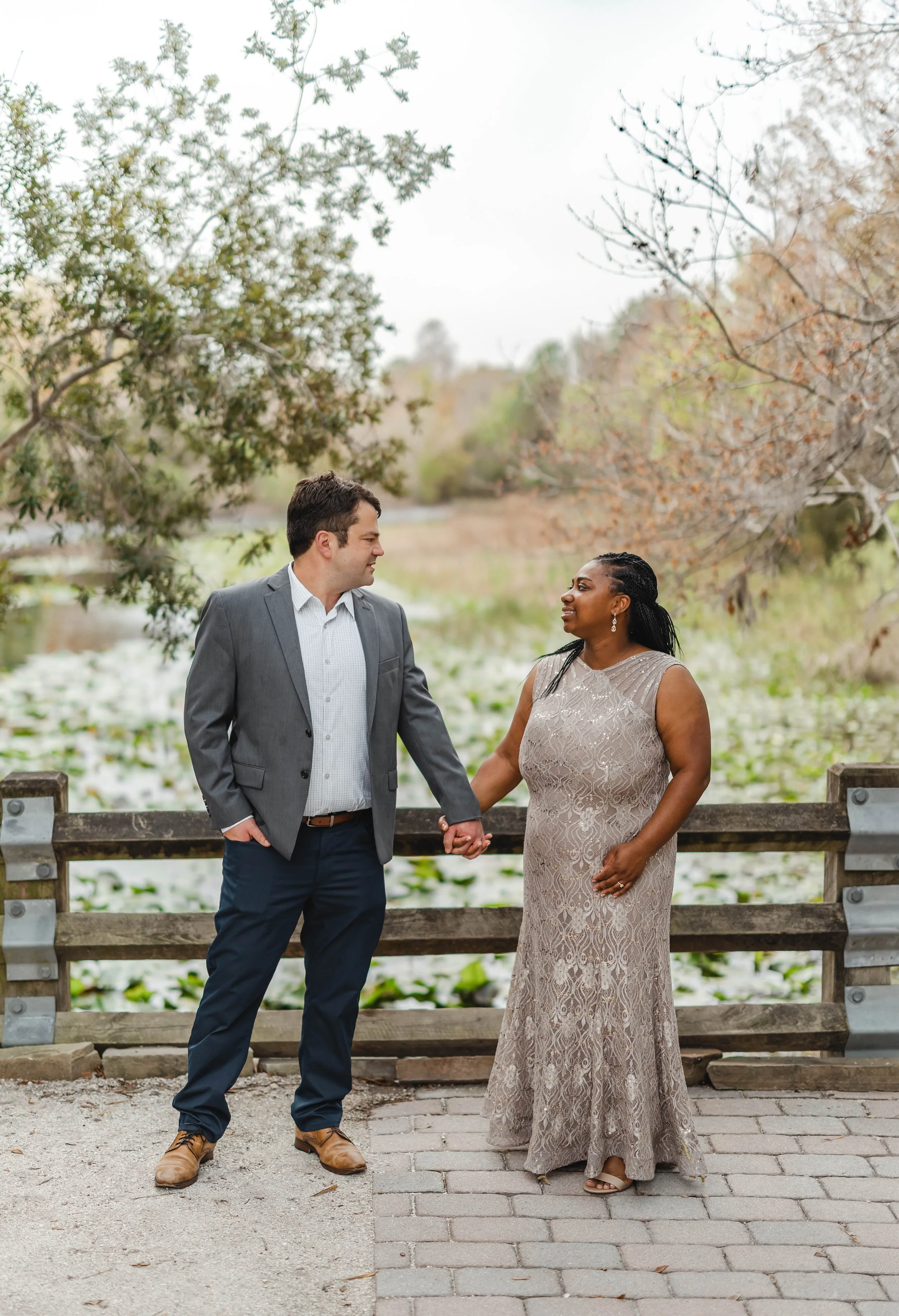 A couple holding hands and looking at each other outdoors near a pond with lily pads, surrounded by trees with some autumn leaves.