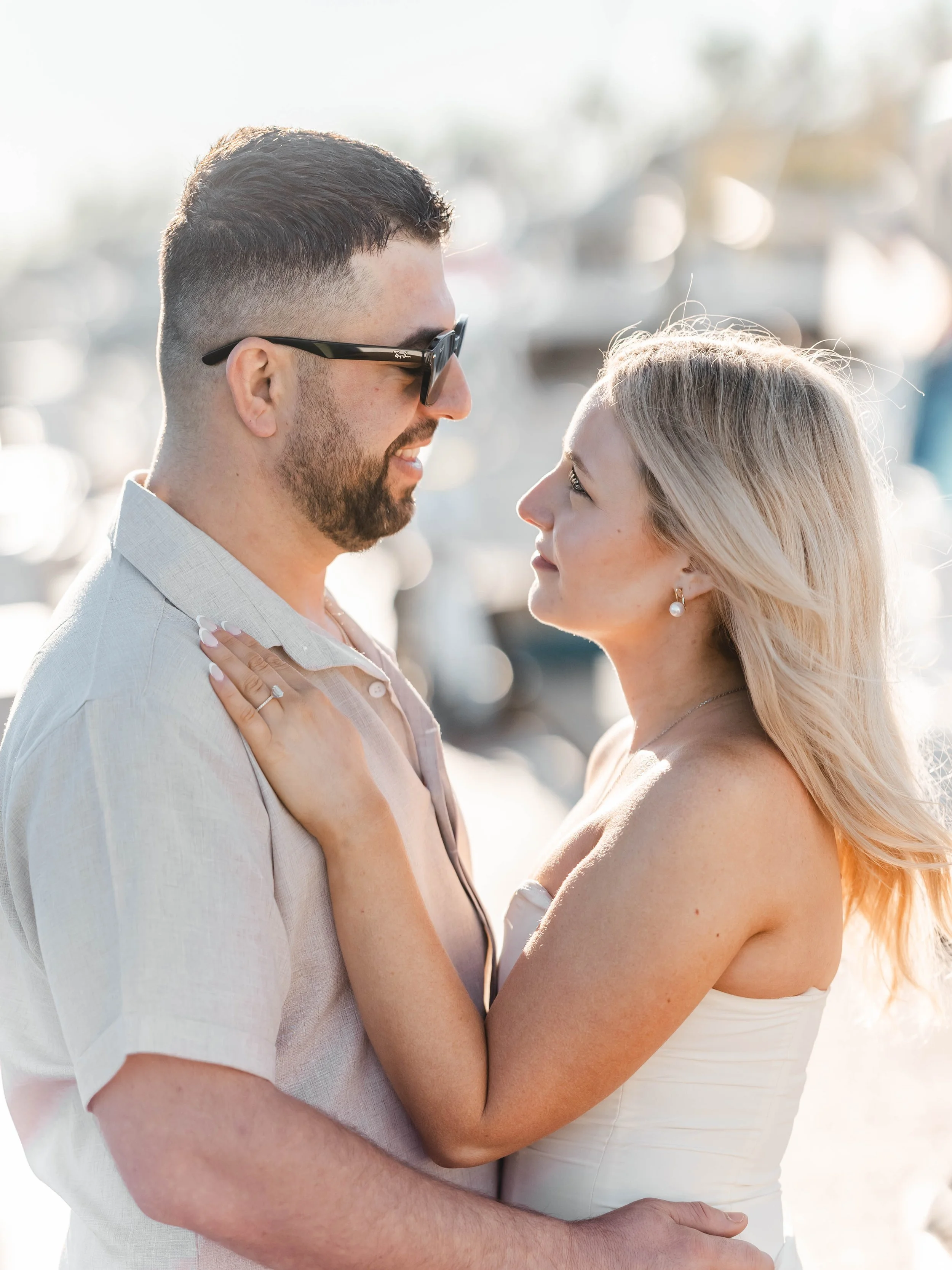 A smiling couple, a man with sunglasses and a woman with blonde hair, are close and looking into each other's eyes at a marina.